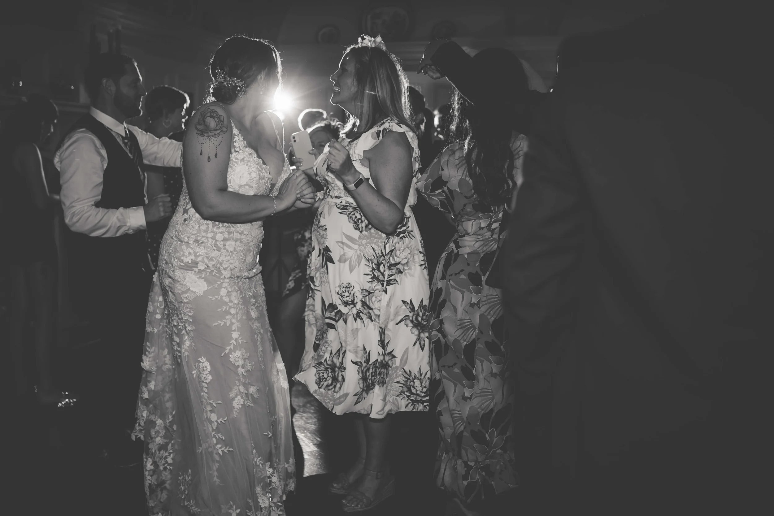 Black and white photo of people dancing and socializing at a wedding reception, with a bride in a lace gown and a woman in a floral dress smiling and holding hands.