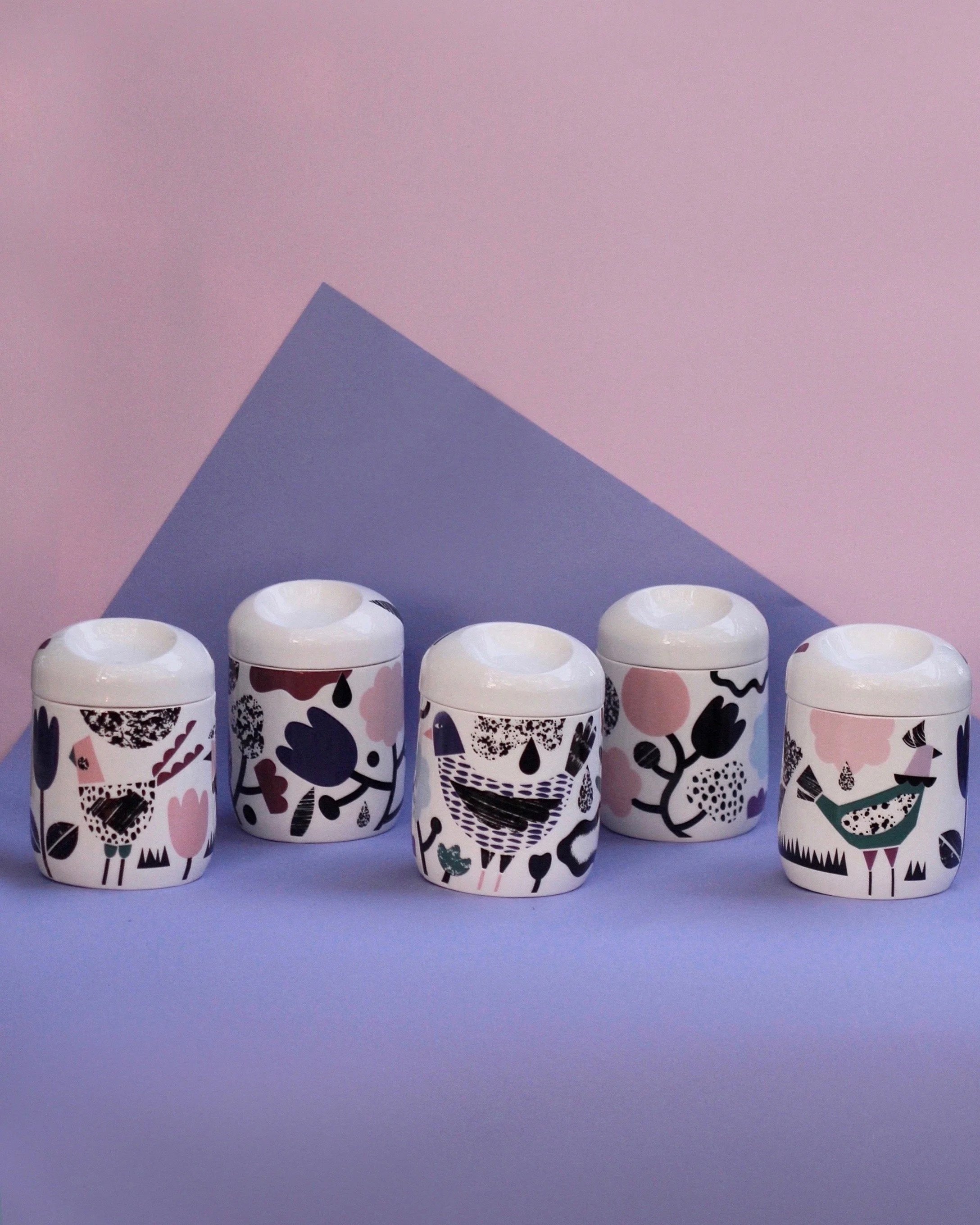 Seven small white containers with colorful, abstract patterns on a purple surface, arranged in two rows, with a pink wall and a purple triangle-shaped object in the background.