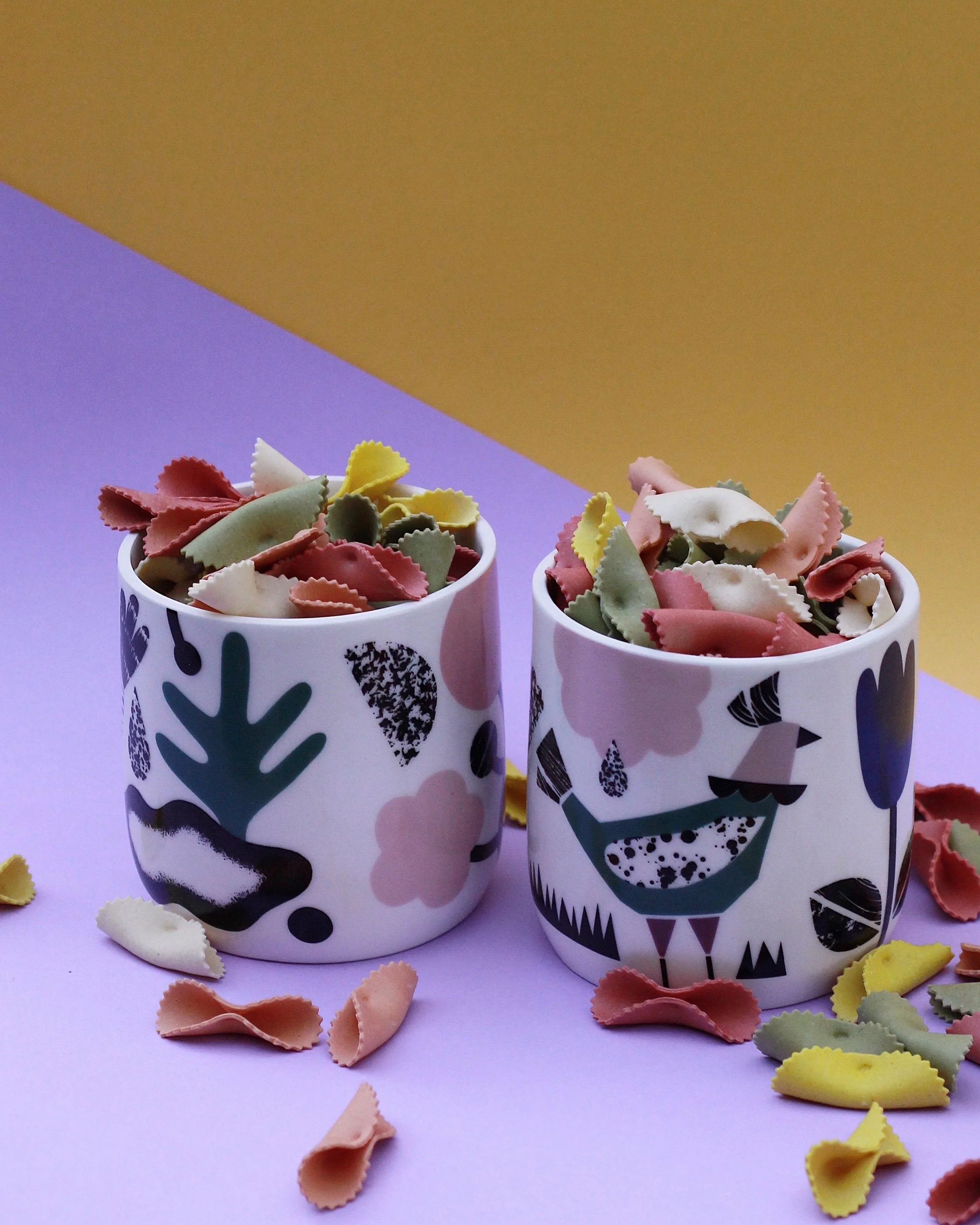 Two cups filled with colorful bowtie pasta-shaped pasta pieces on a purple surface, with additional pasta scattered around.