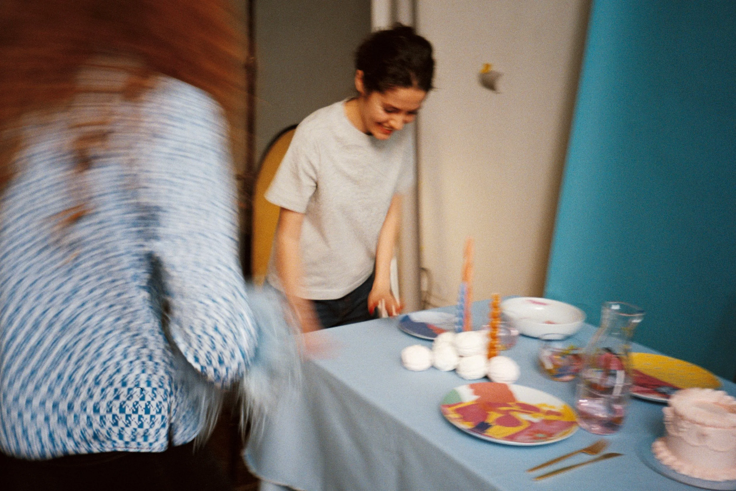 A woman with short dark hair smiling at a birthday celebration, standing next to a table decorated with party supplies, cake, and balloons.