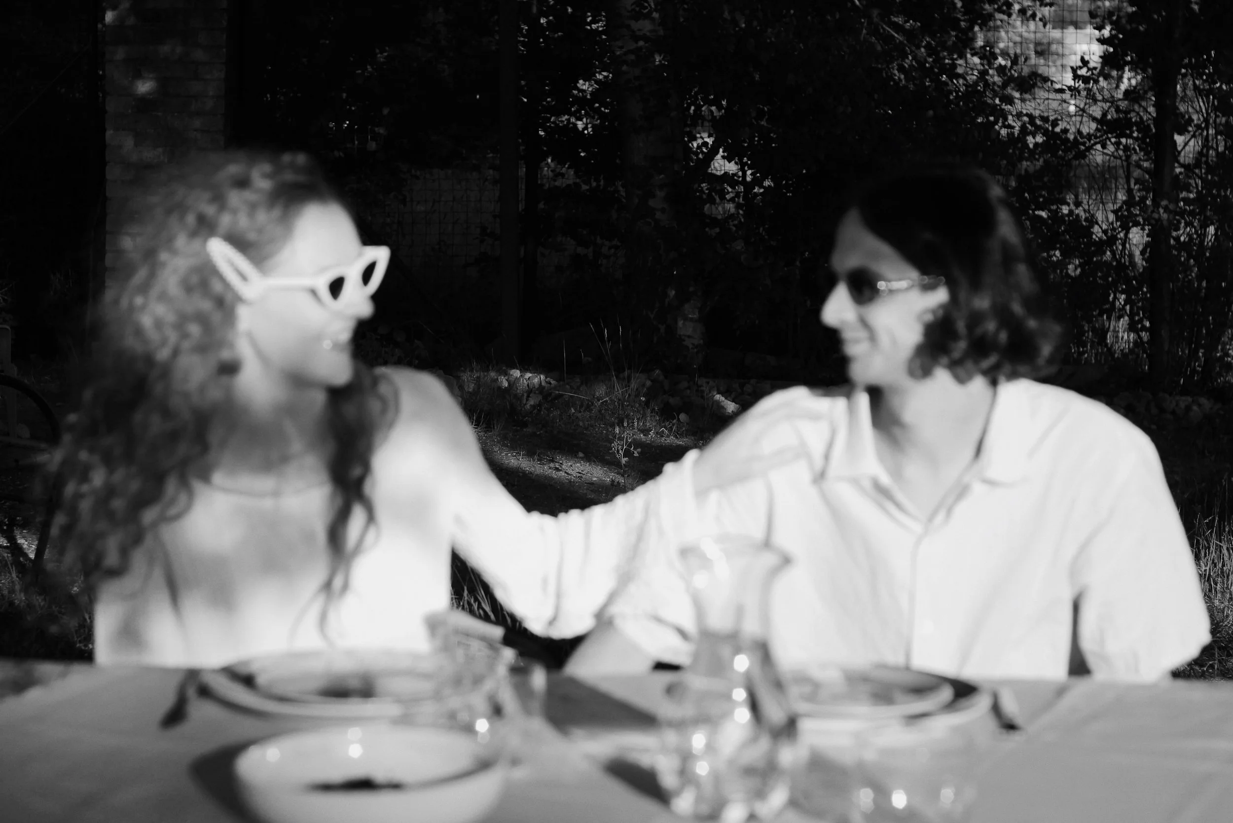 Two women with glasses smiling and talking outdoors at night while sitting at a table.