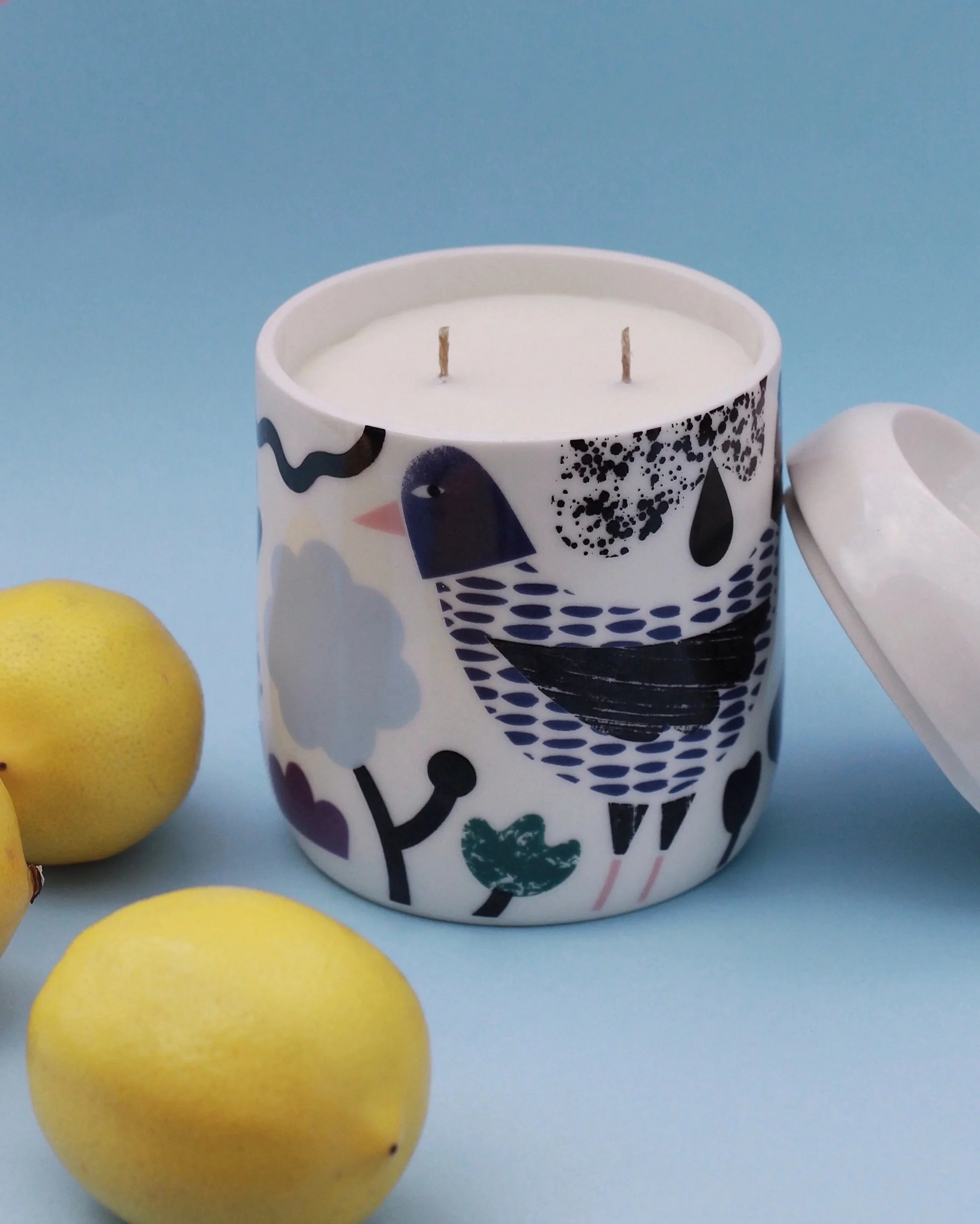 A decorative white candle in a colorful bird illustrated mug with two wicks inside, and three yellow lemons partially visible in the foreground with a light blue background.