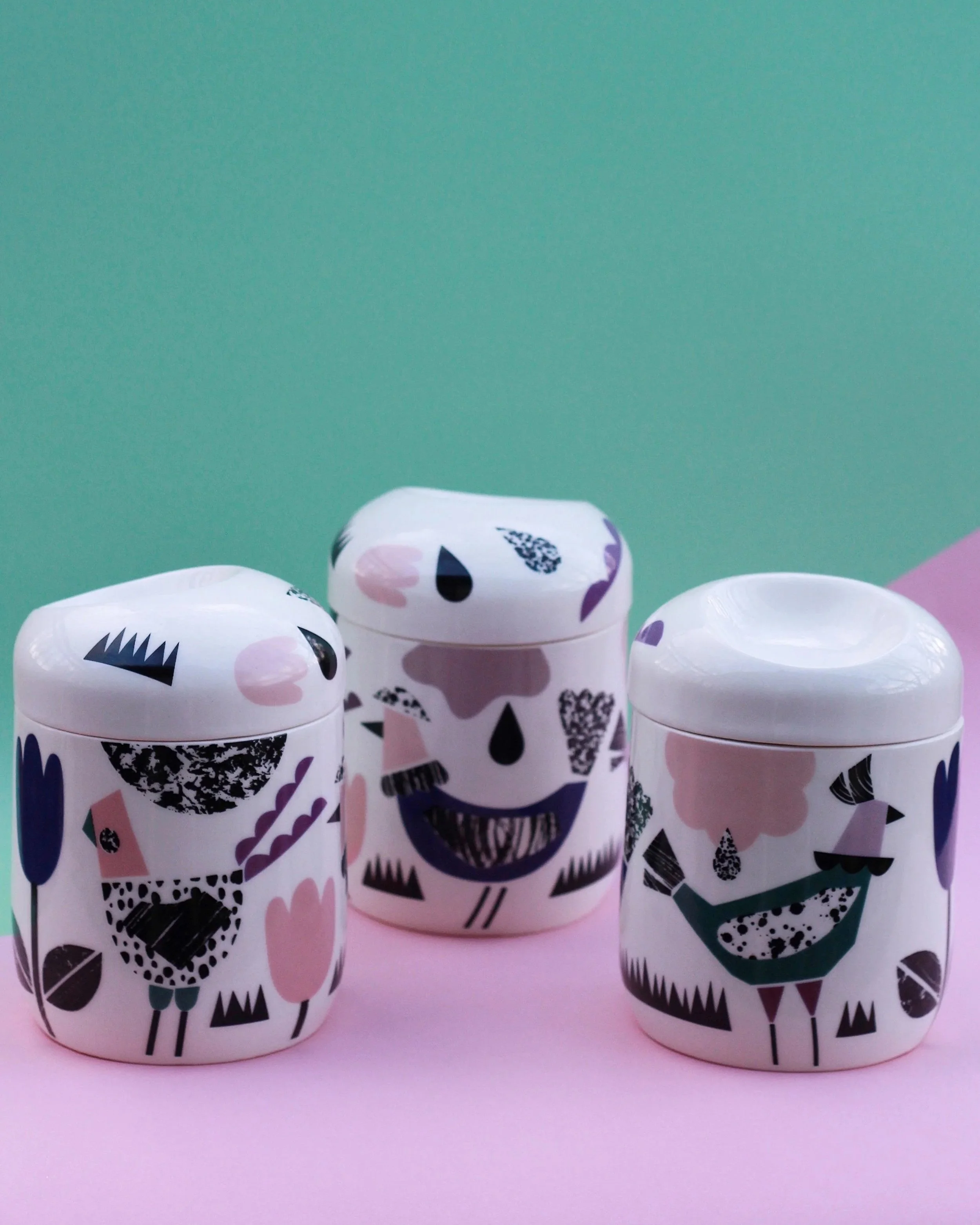 Four cans decorated with colorful abstract birds, placed on a pink surface with a green background.
