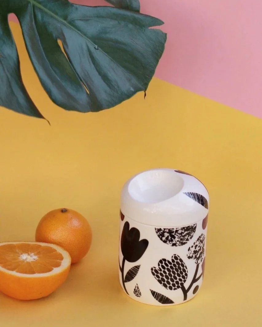 A white ceramic jar with black floral patterns, a whole orange, a halved orange showing the inside, and a large green leaf on a yellow surface against a pink and yellow background.