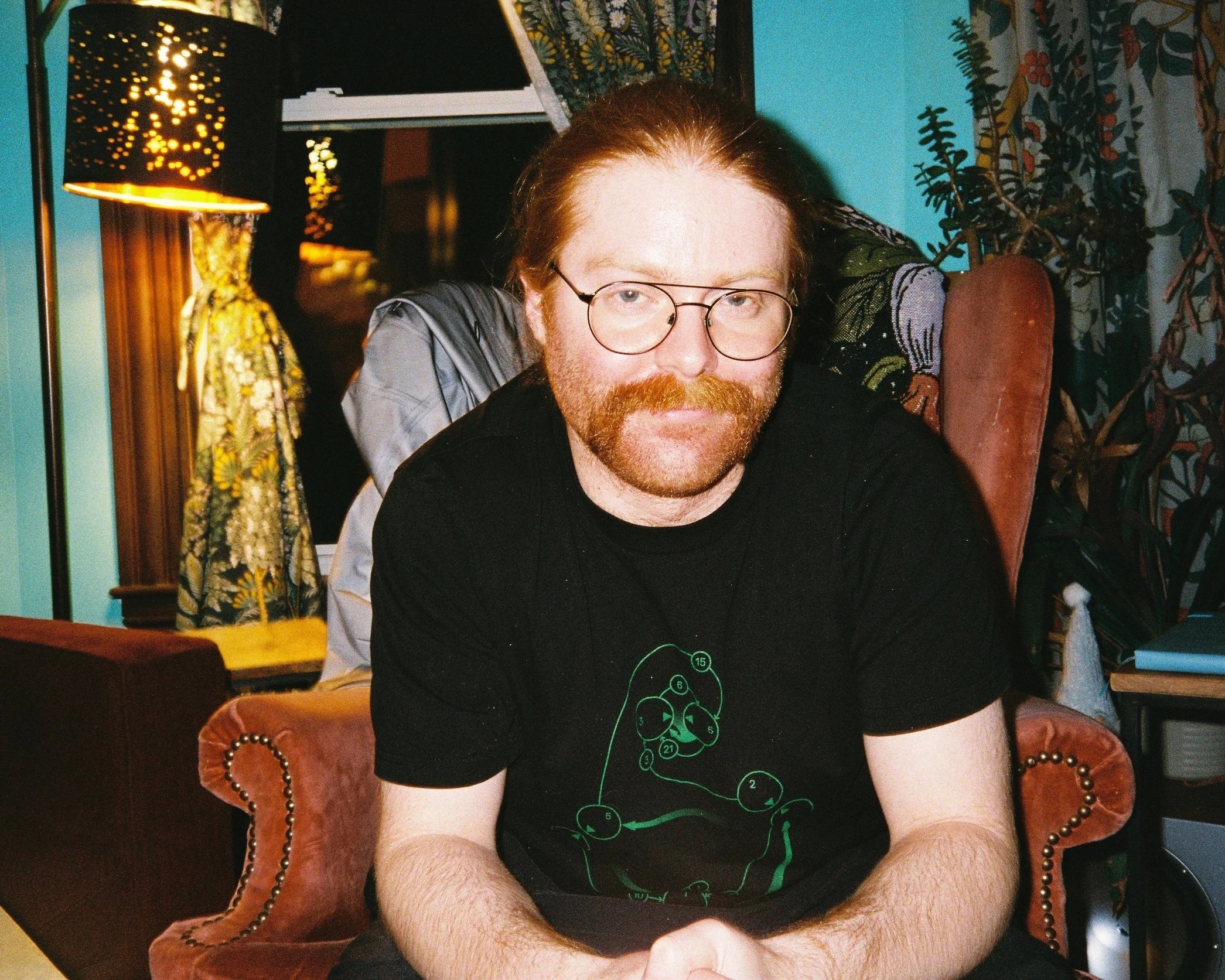 A man with red hair and beard wearing glasses and a black shirt with a numogram on it, sitting in a vintage-style room with patterned curtains, a wooden furniture piece, a lamp, and a teal-colored wall.