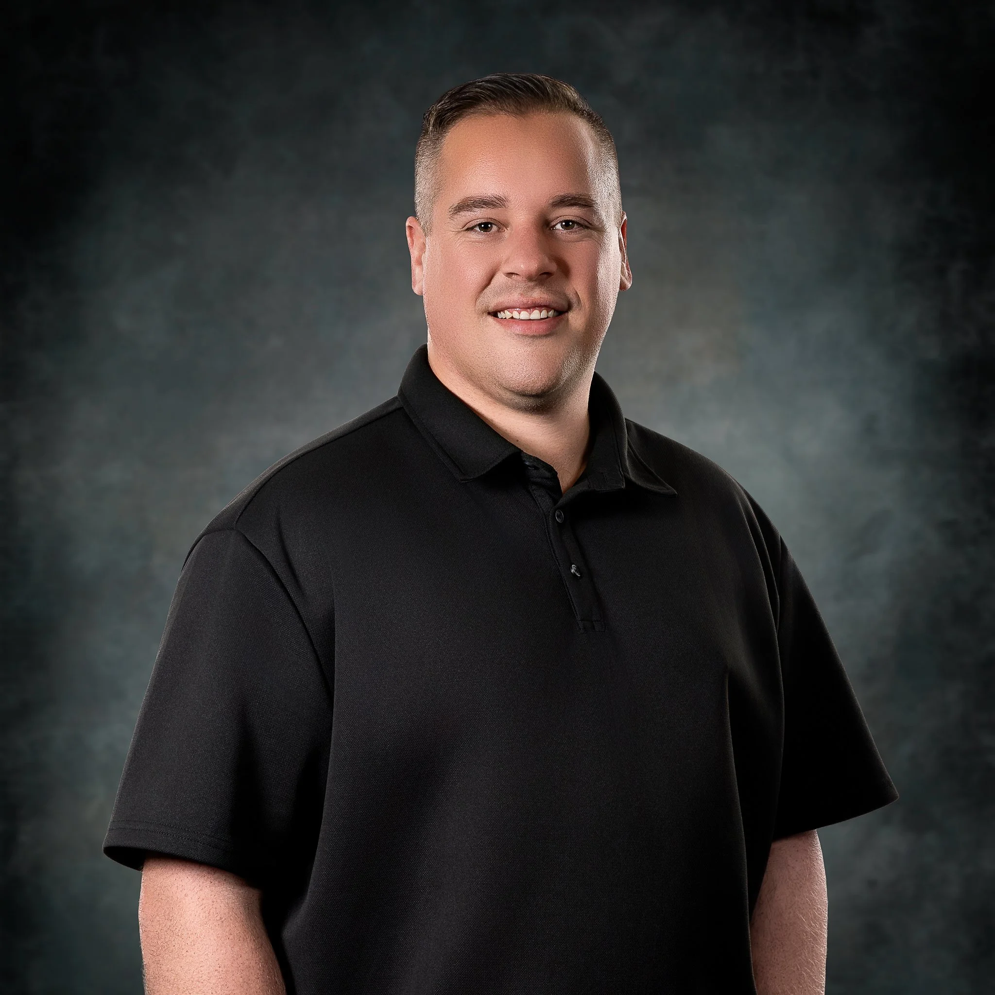Close-up of a young man with short brown hair, smiling, wearing a black polo shirt, against a dark, textured background.