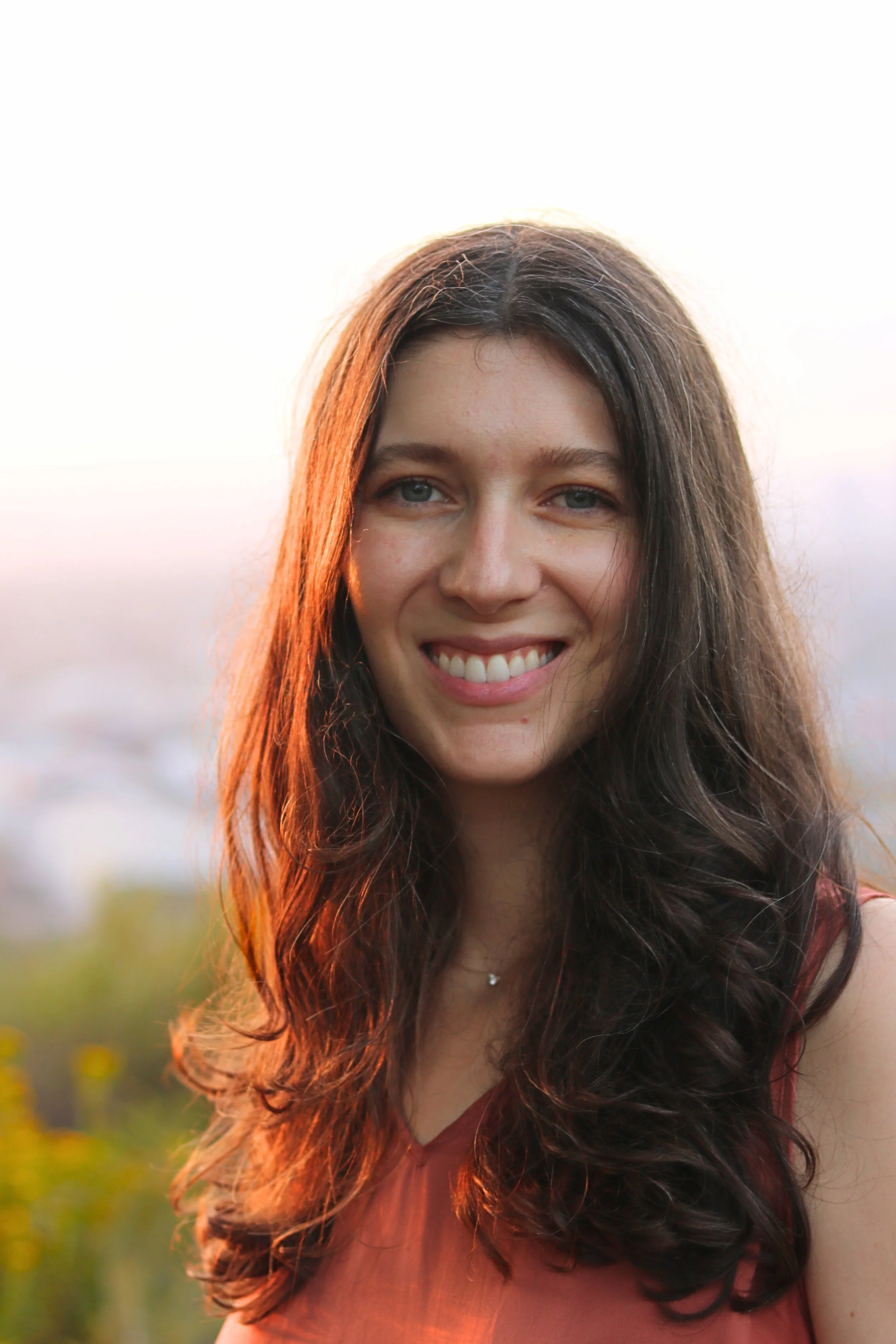 Close-up of a smiling woman with long wavy brown hair, outdoors at sunset with blurred greenery in the background.