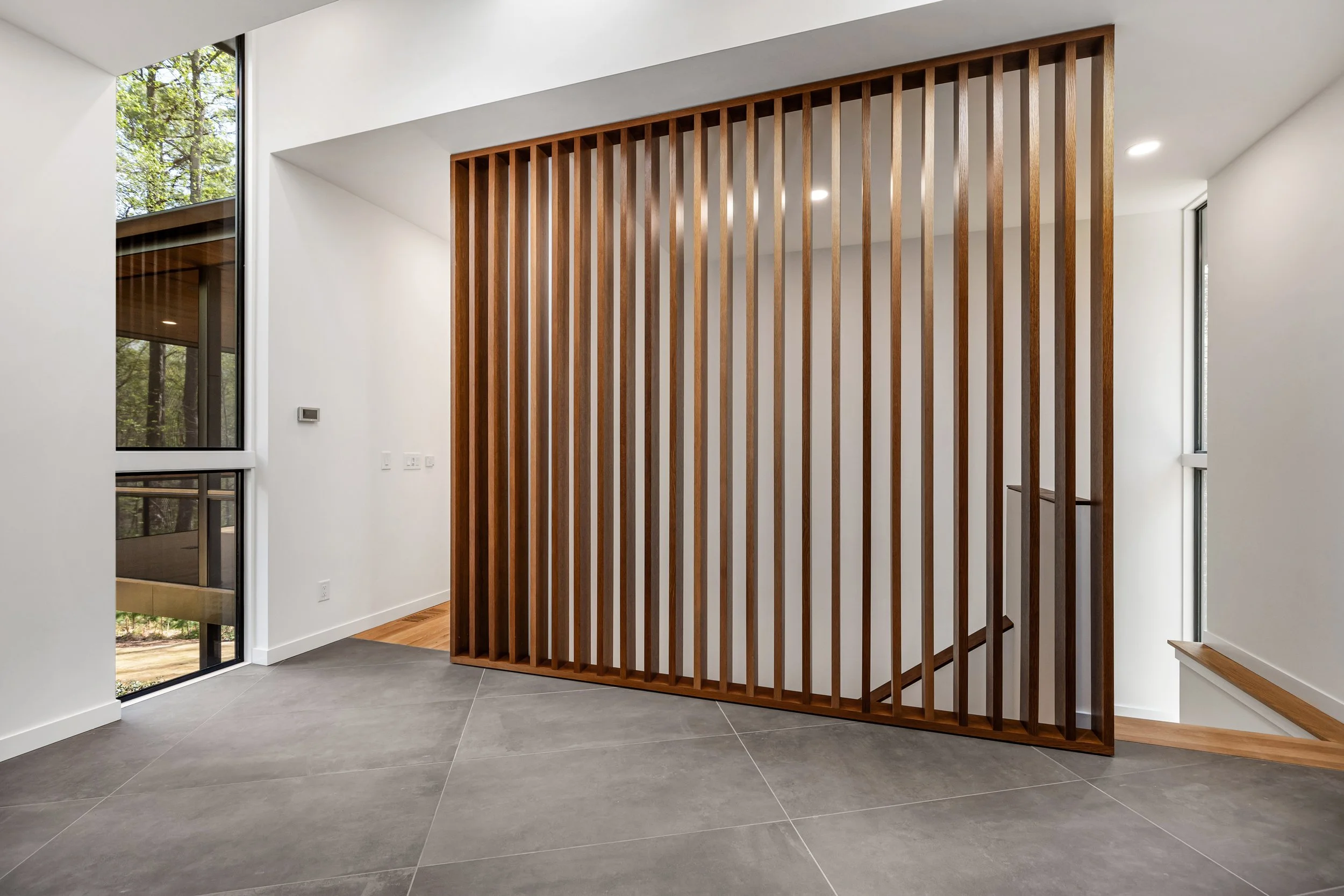  The foyer is lit by a dramatic slot of light from left and above. The stair down is screened by an open wall of heavy timbers. 