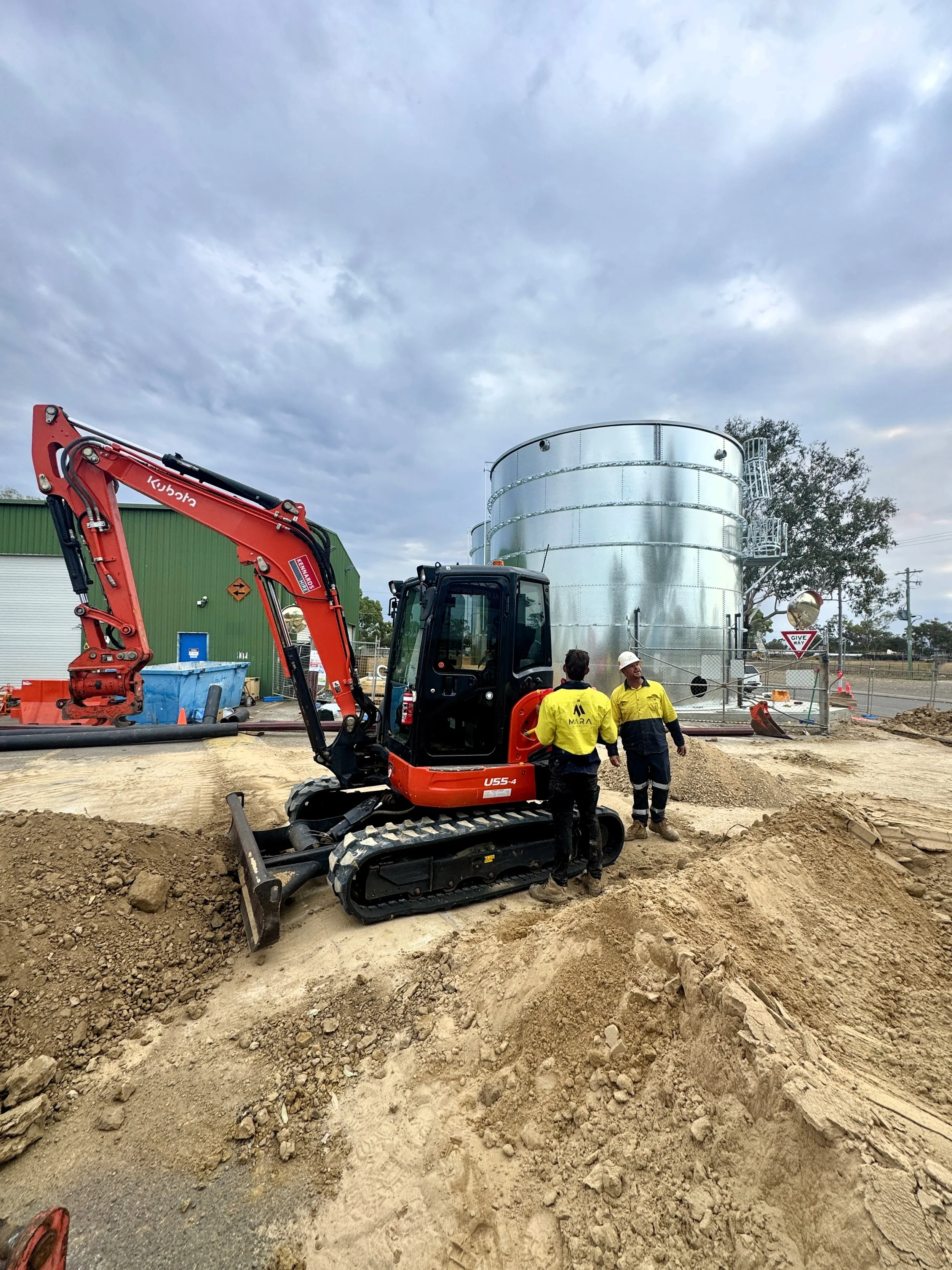 Construction site with workers and heavy machinery, including a small excavator, in front of large metallic tank and green building, under cloudy sky.