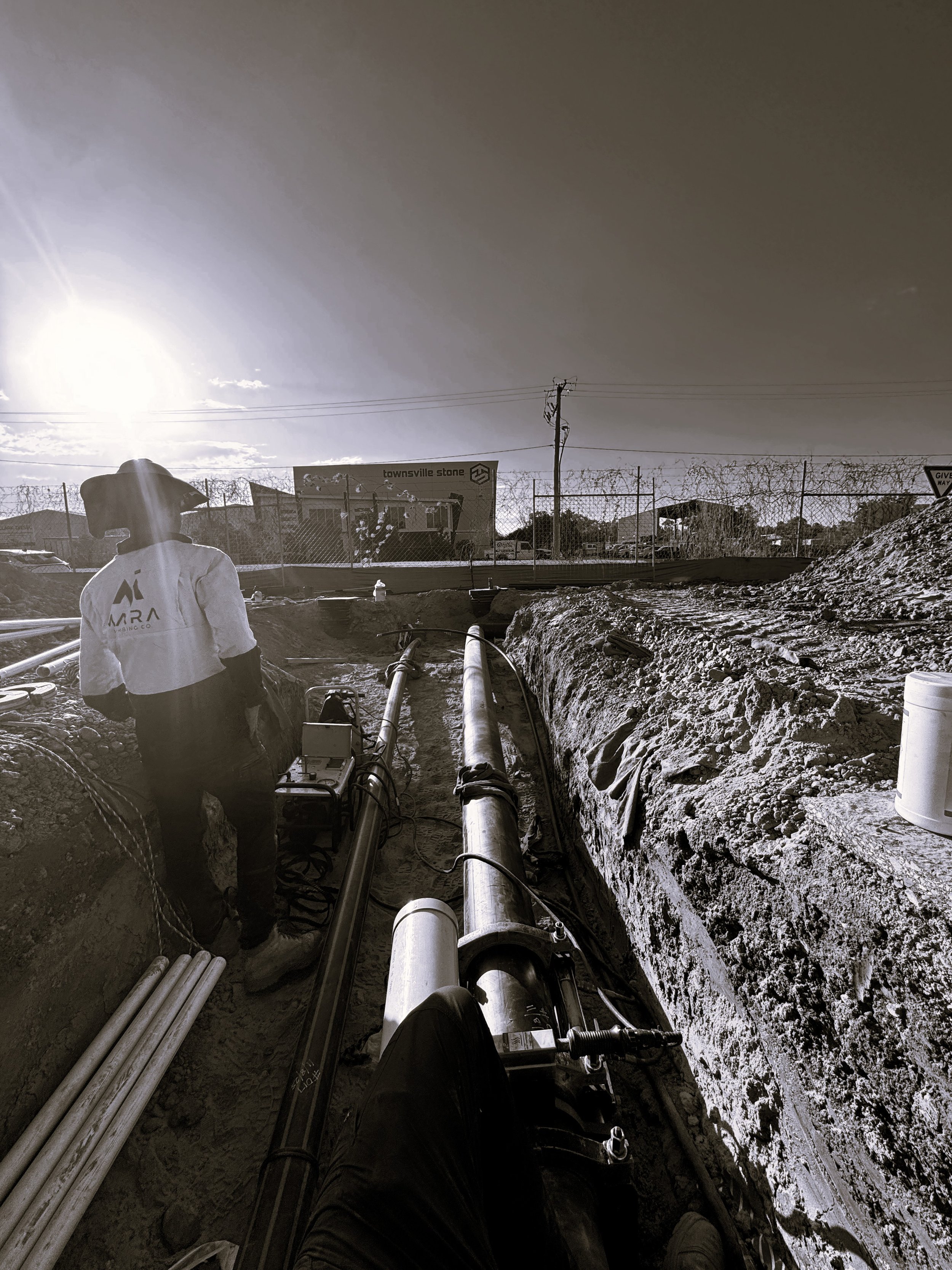 A person standing on a construction site near pipelines and pipes underground, with a chain-link fence and a building labeled 'Townsville Stone' in the background, under a bright sun in the sky.