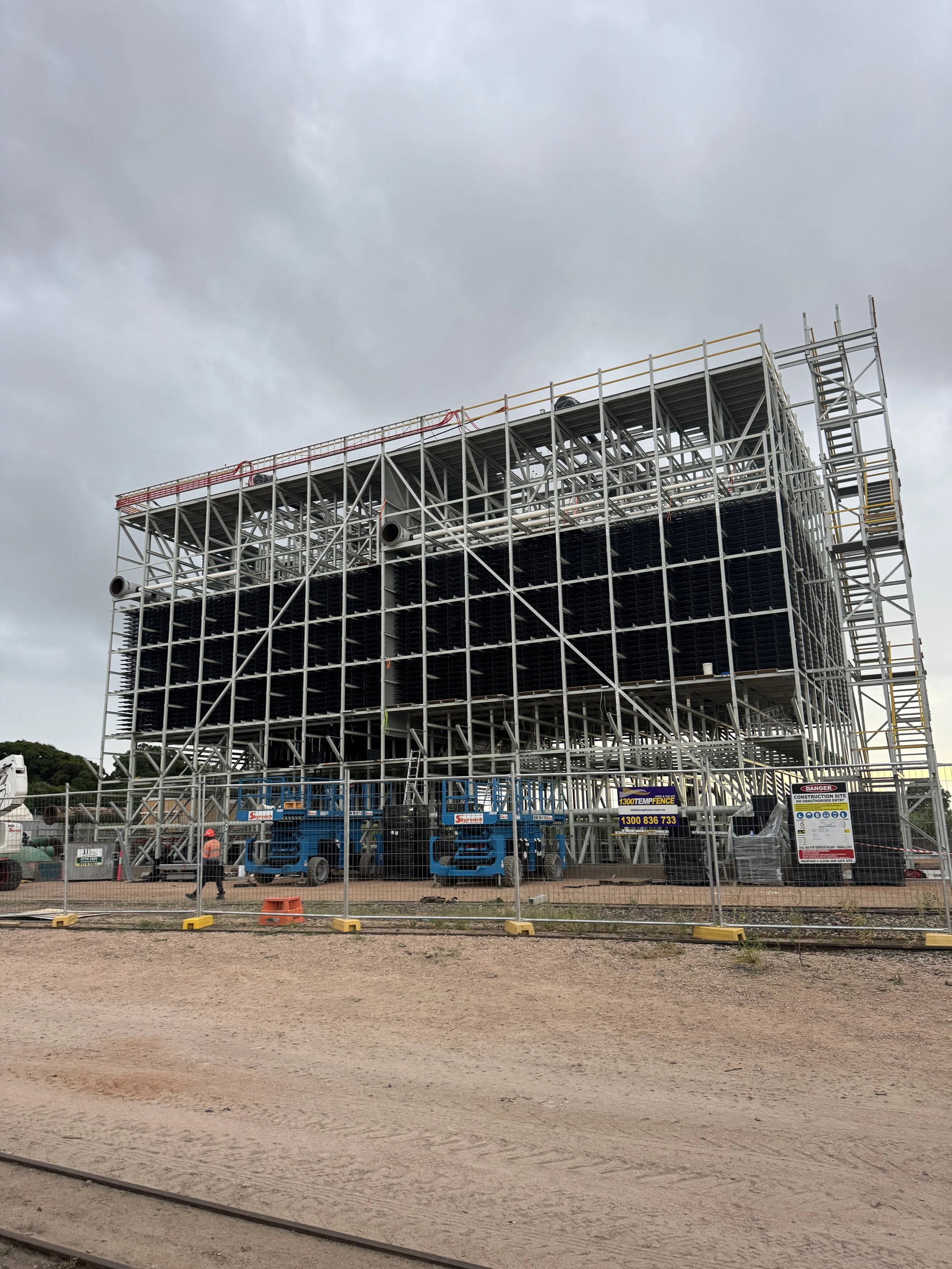 A construction site with a large steel framework for a building, surrounded by a fence, with workers and construction vehicles present, and cloudy sky overhead.