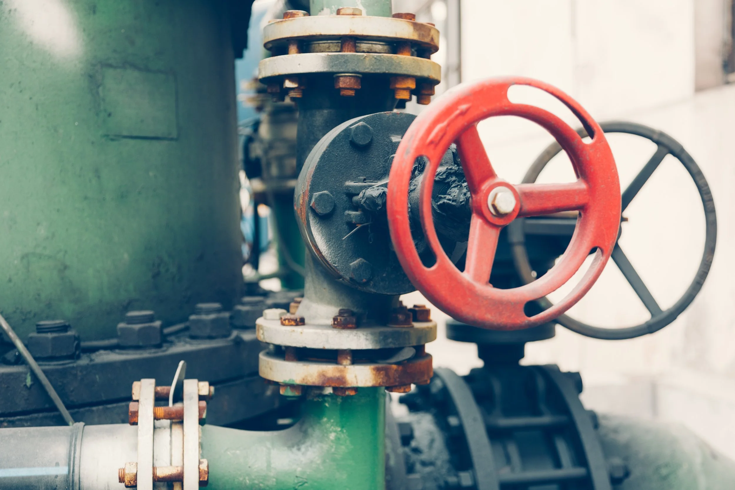 Close-up of industrial pipes and valves, featuring a red wheel valve on a black pipe, with green and rusted metallic components.