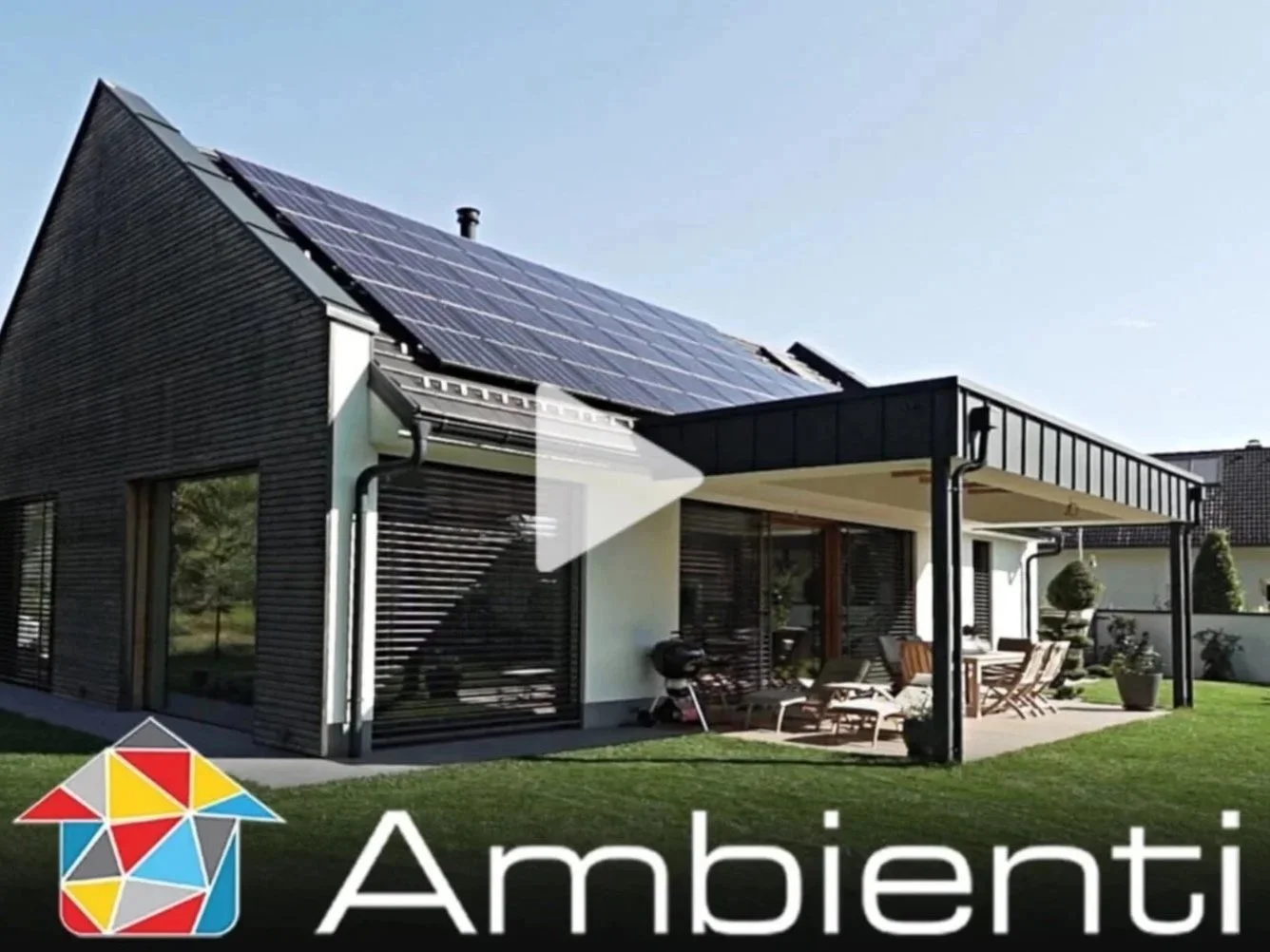 Modern house with solar panels on the roof, large windows, a patio with outdoor furniture, and a well-maintained lawn, with the word 'Ambienti' and a colorful house logo at the bottom.