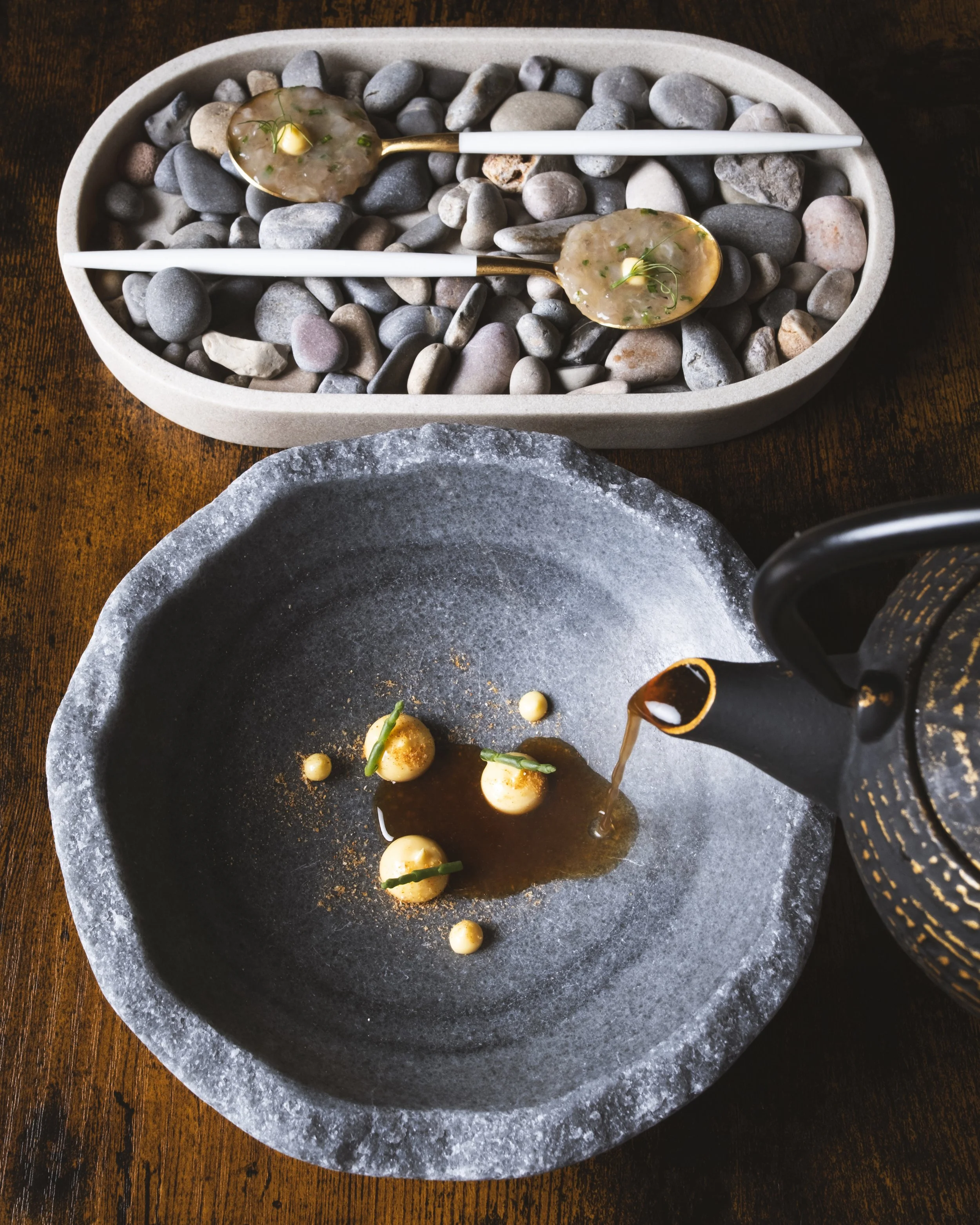 Tazones de sopa con caldo oscuro y esferas pequeñas decorativas, uno siendo servido con un vertido de caldo, sobre una mesa de madera, con piedras decorativas en un recipiente al fondo.