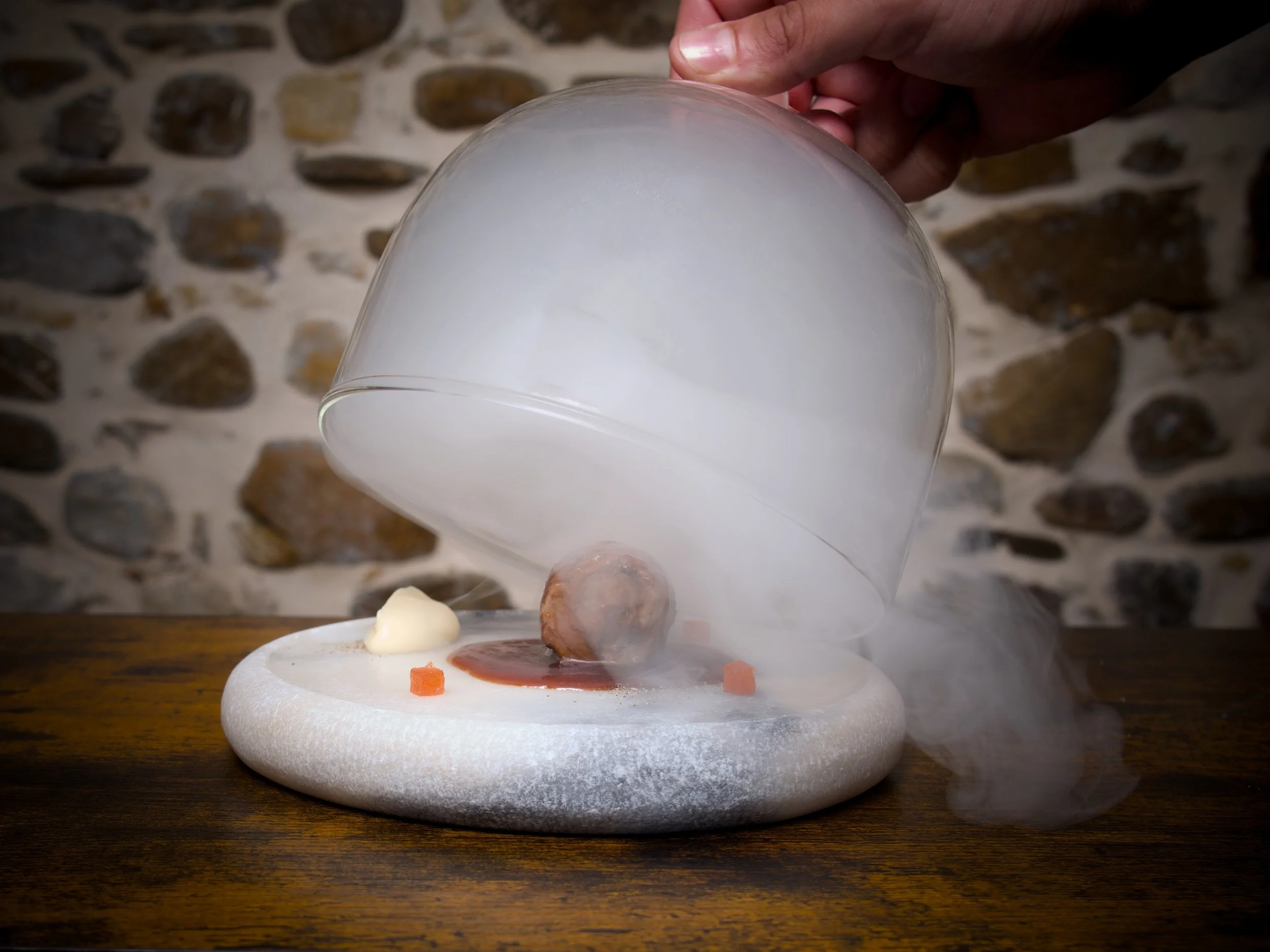 Presentación de plato gourmet con humo, carne, salsas y complementos en mesa de madera.