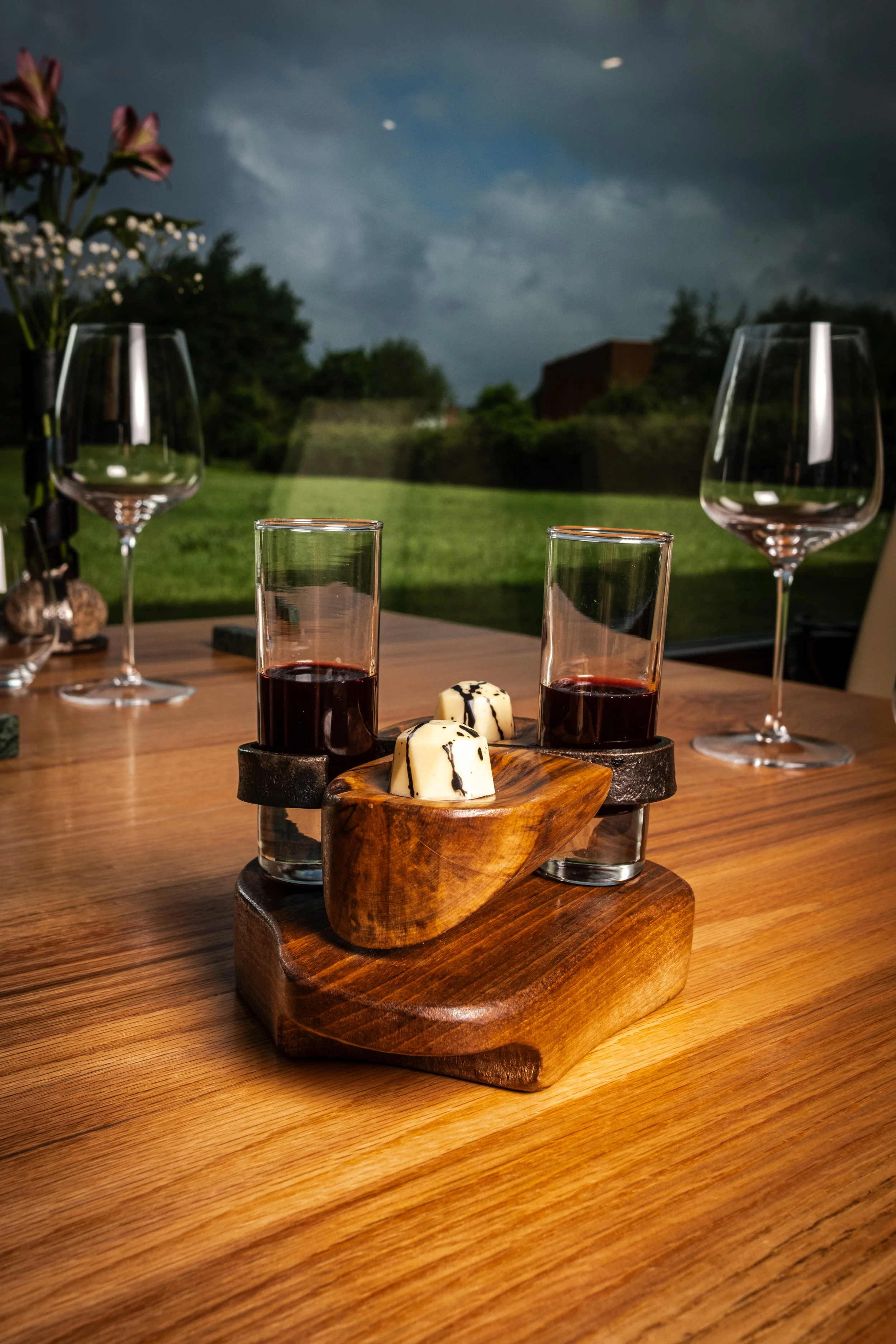 Mesa con dos copas de vino tintóny dos vasos con vino tinto, acompañados de queso en una bandeja de madera, con un fondo de paisaje exterior con cielo nublado.