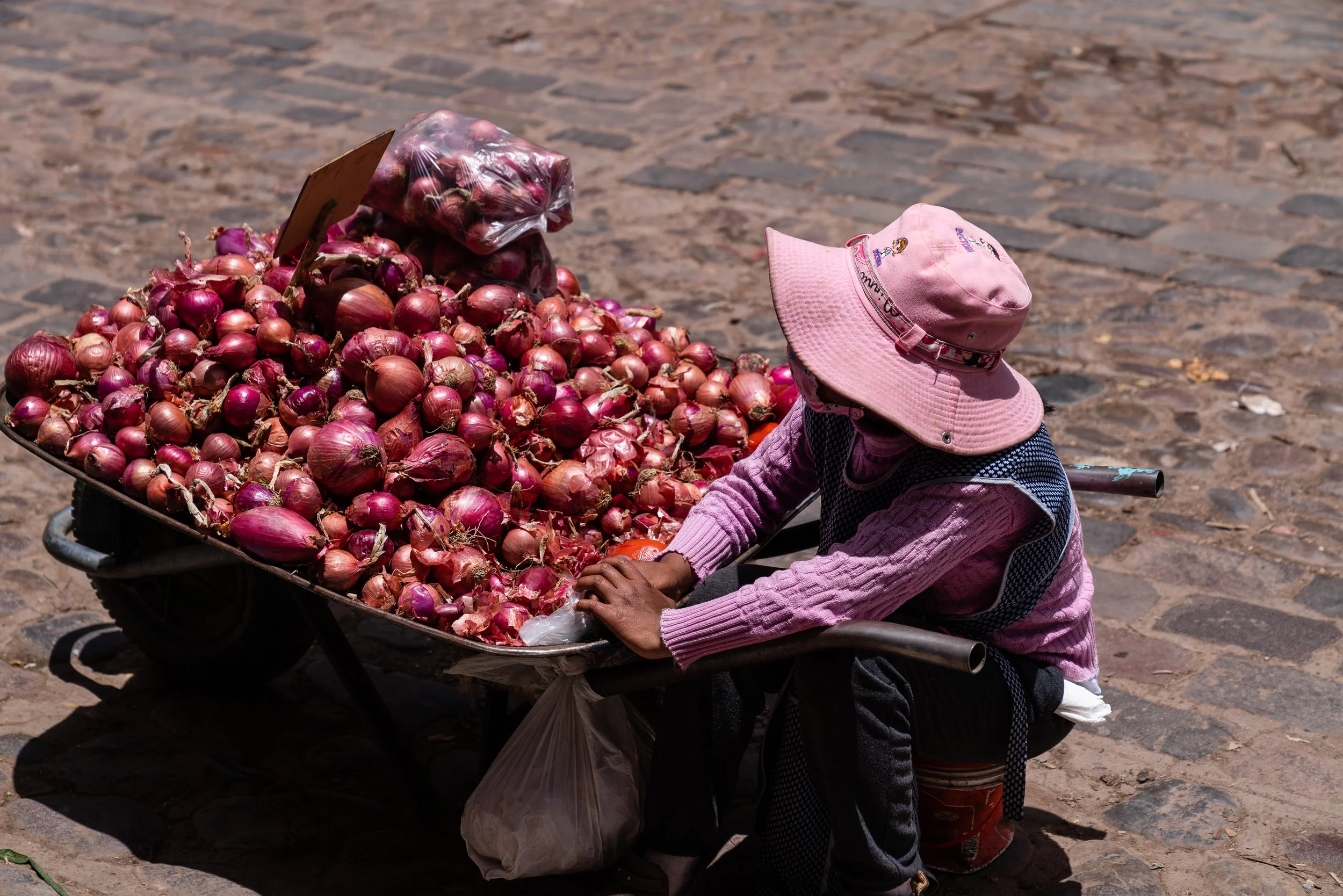 Person wearing pink clothes and hat sitting by a wheelbarrow full of red onions on a cobblestone street.