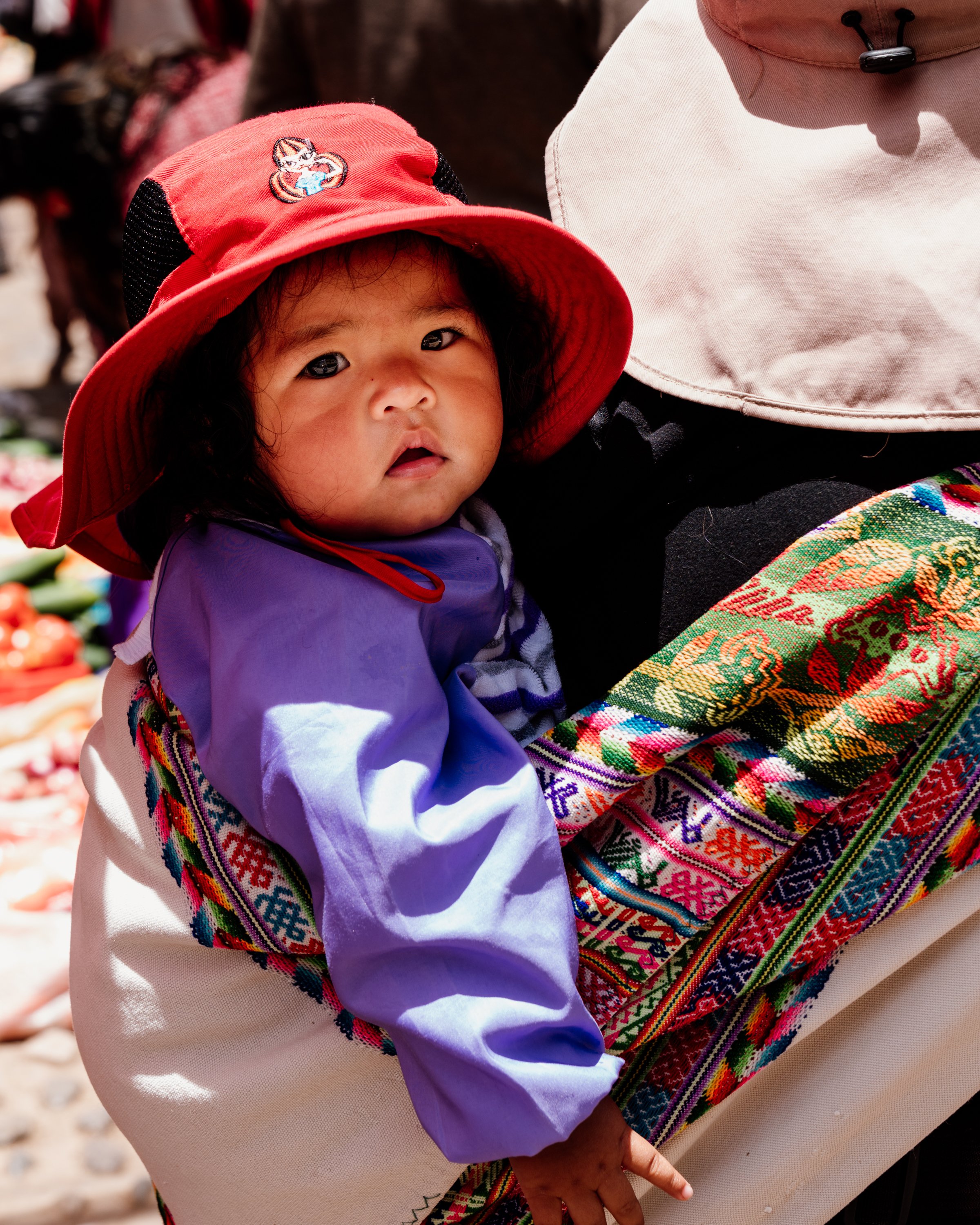 Child wearing a red hat and blue shirt, carried in colorful woven fabric.