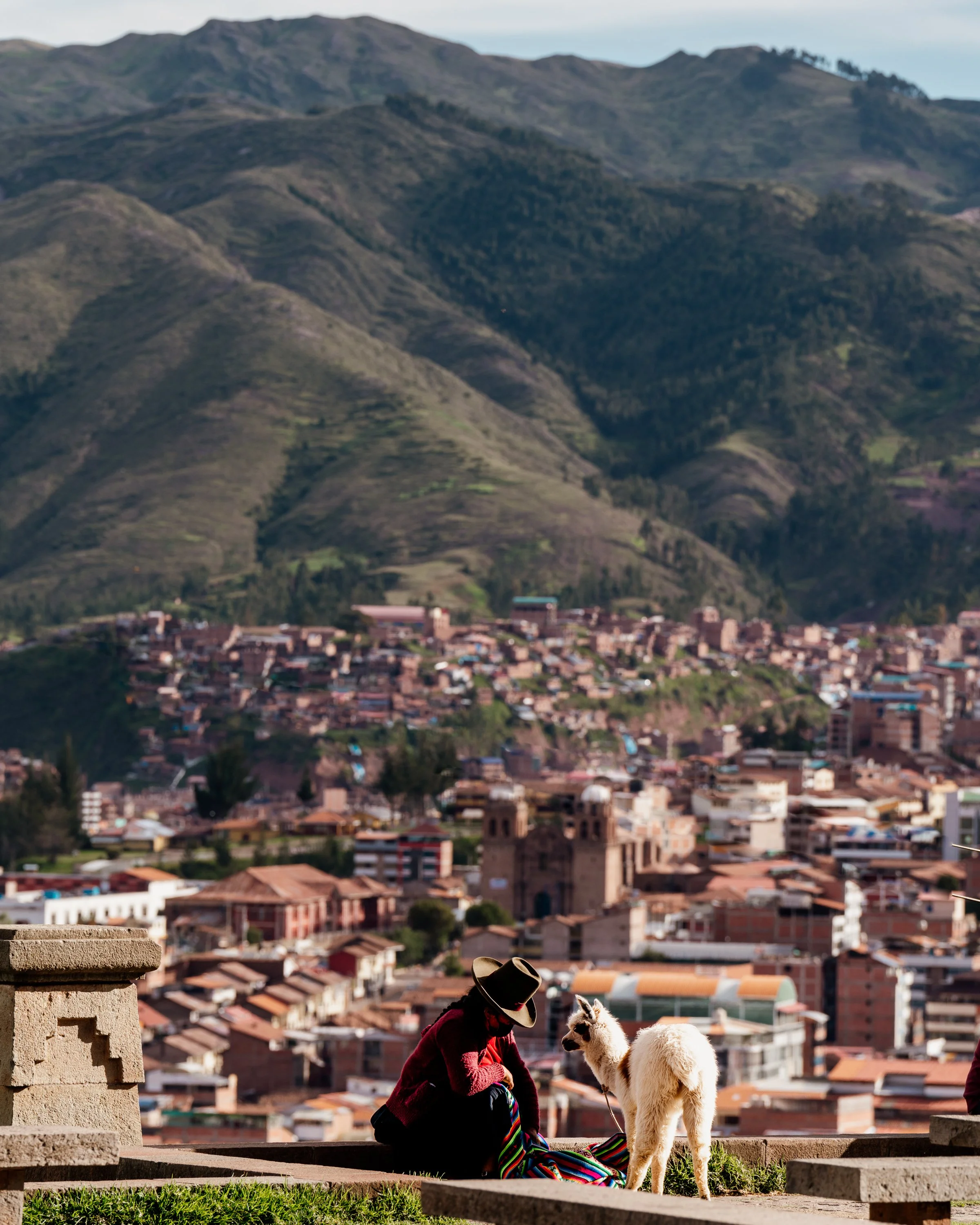 Person in traditional clothing with a llama in Cusco, Peru, overlooking cityscape and Andean mountains.