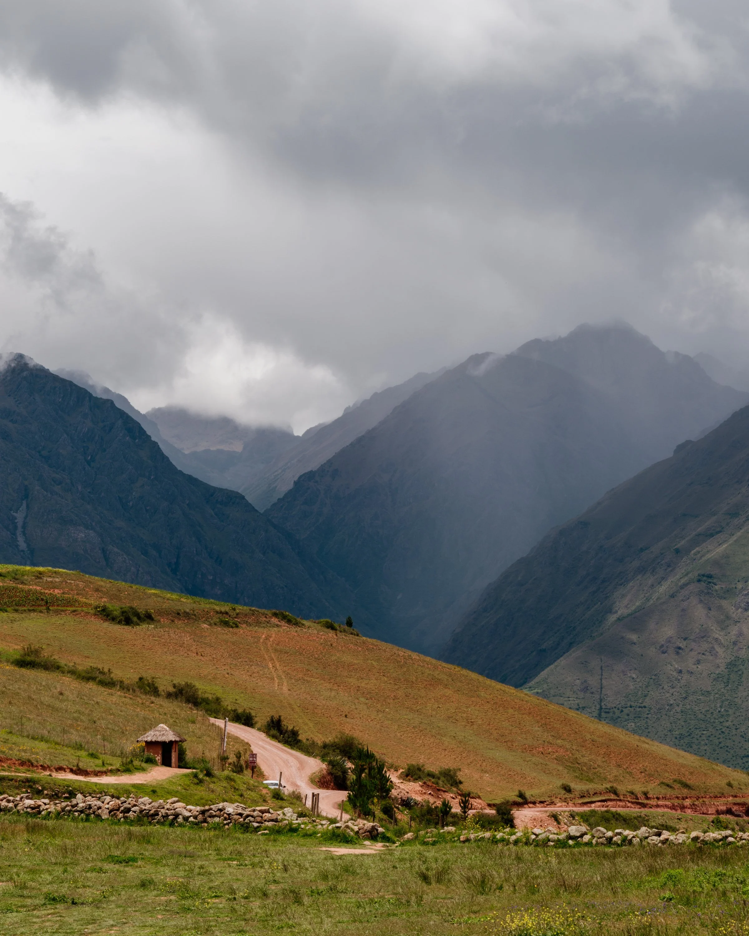 A scenic landscape with green hills, a dirt road, and a small thatched-roof hut in the foreground, surrounded by mountains under a cloudy sky.