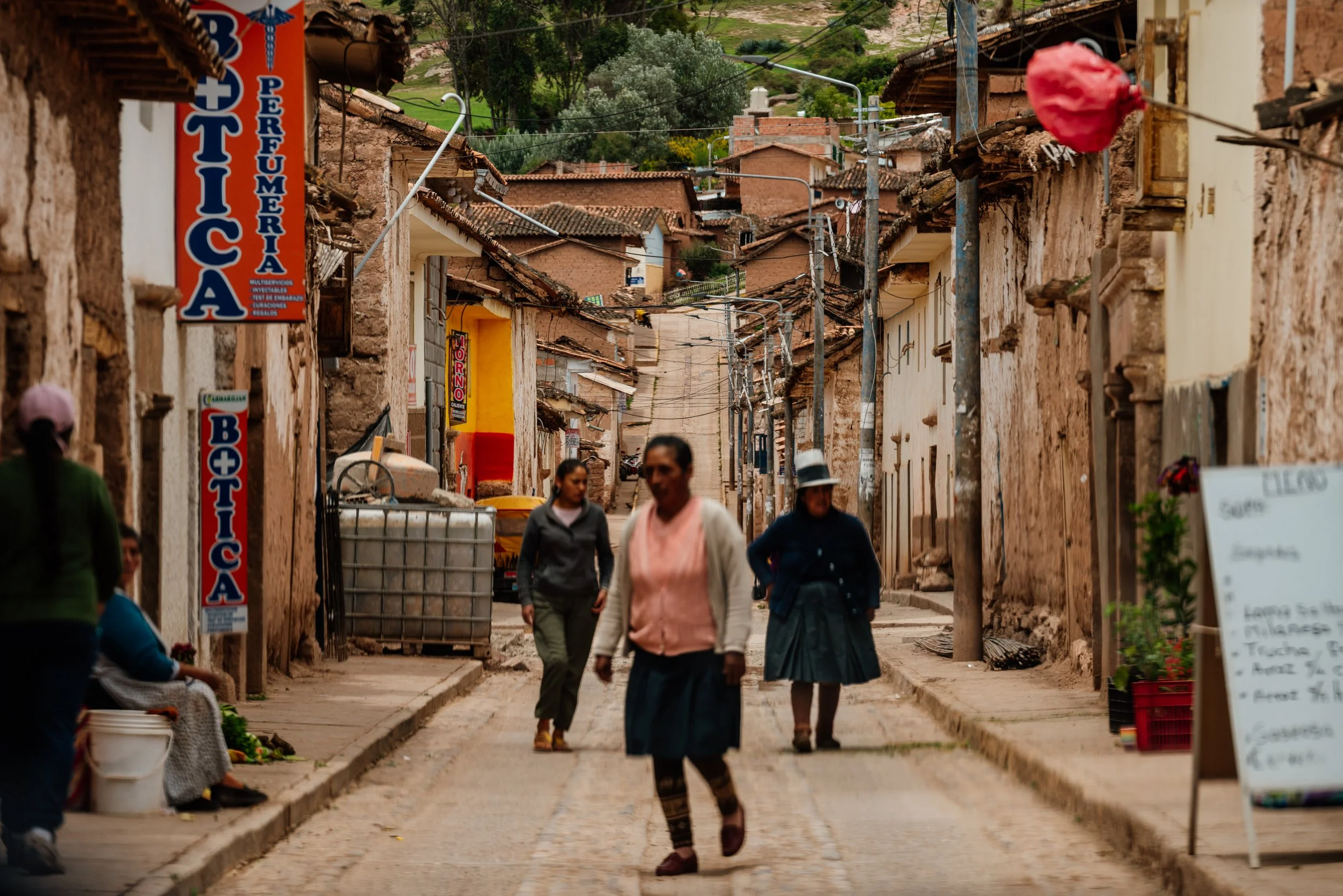 A narrow street in a small village with people walking, adobe buildings, and a pharmacy sign in Spanish. There are power lines and hills in the background.