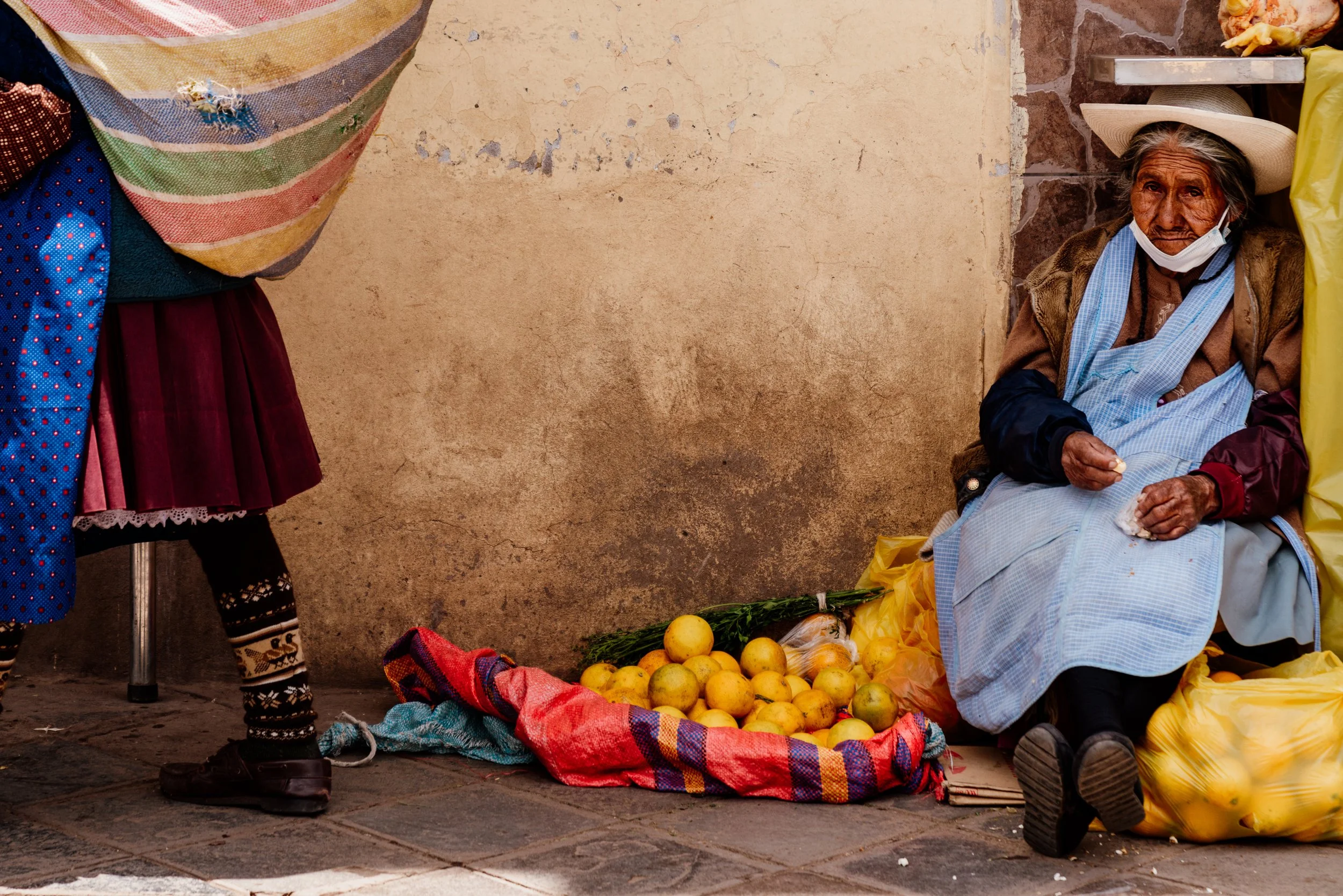 Elderly woman sitting beside a pile of oranges on a sidewalk, wearing a hat and apron; another person walks by carrying a striped bag.