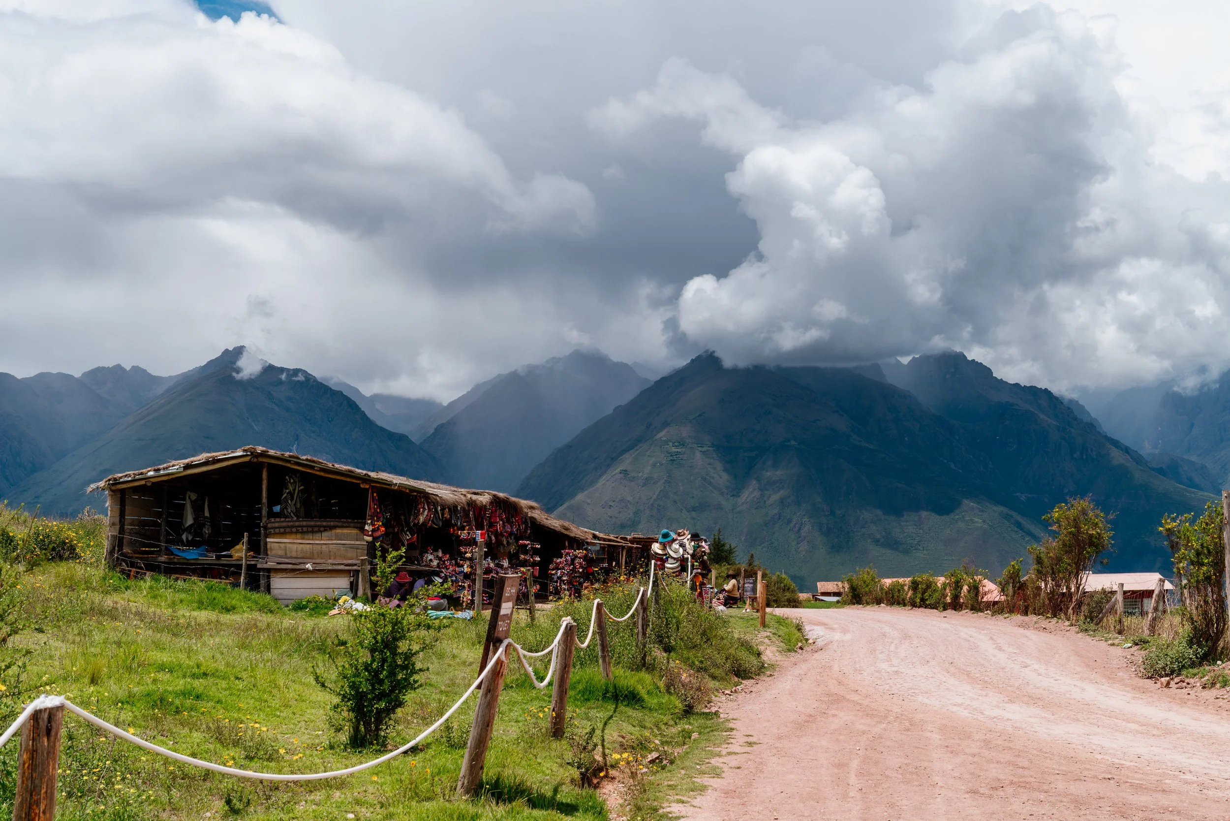 Rustic building with a thatched roof surrounded by green grass, with dramatic mountainous landscape and cloudy sky in the background.