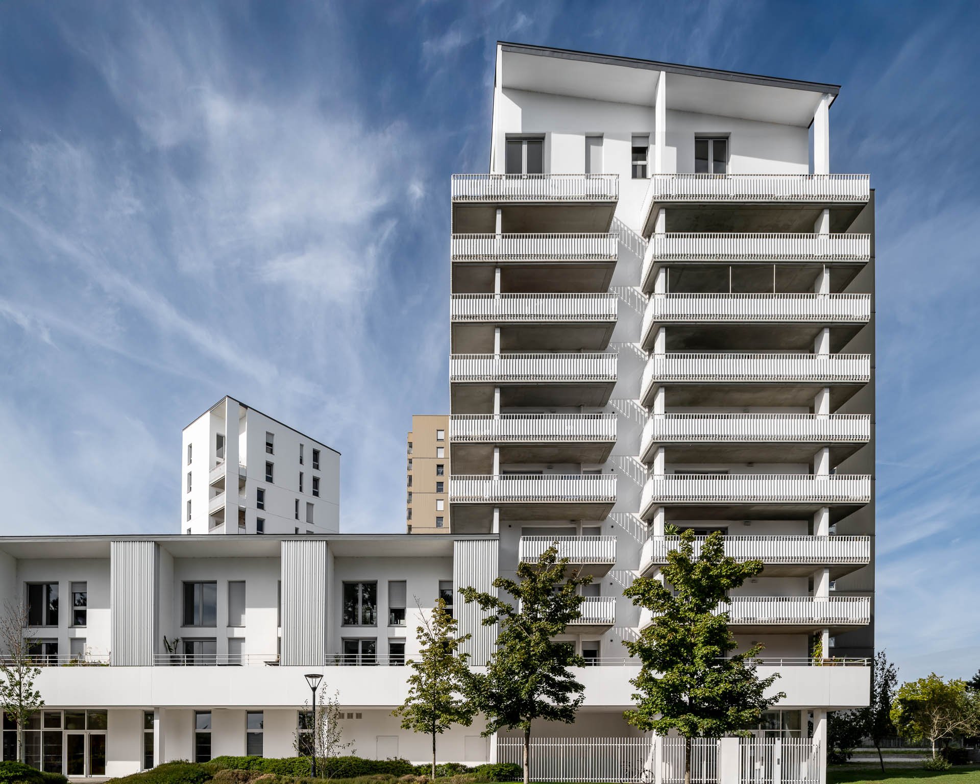 Bâtiment résidentiel moderne blanc avec plusieurs balcons et arbres devant, ciel bleu avec nuages légers.