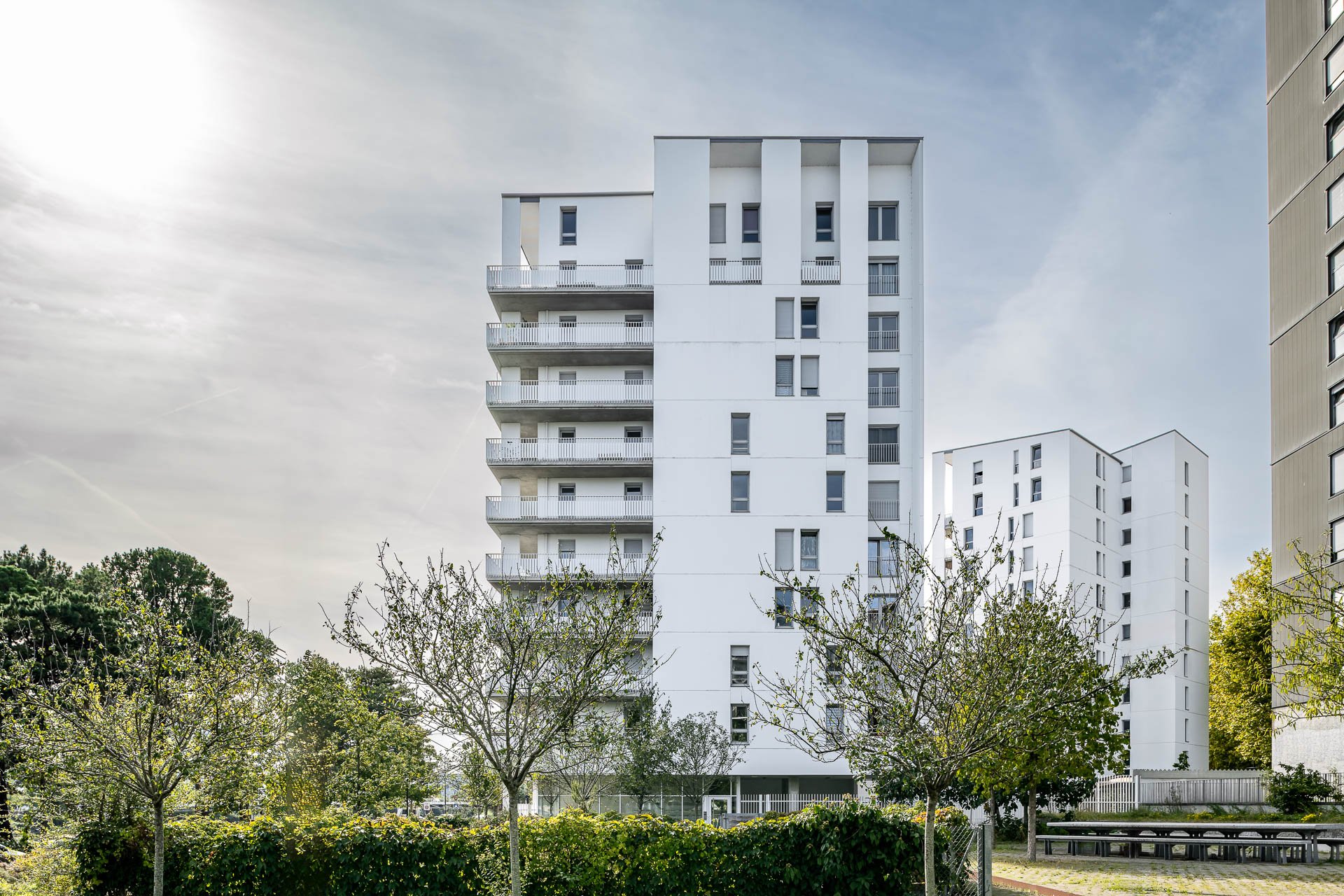 immeuble blanc moderne avec plusieurs étages, balcon, et fenêtres, entouré d'arbres et de végétation, sous un ciel partiellement nuageux.