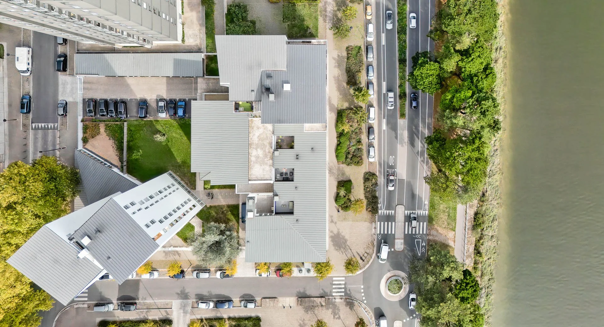 Vue aérienne montrant un quartier résidentiel avec plusieurs immeubles, des voitures stationnées, des arbres verts et une rivière à droite.