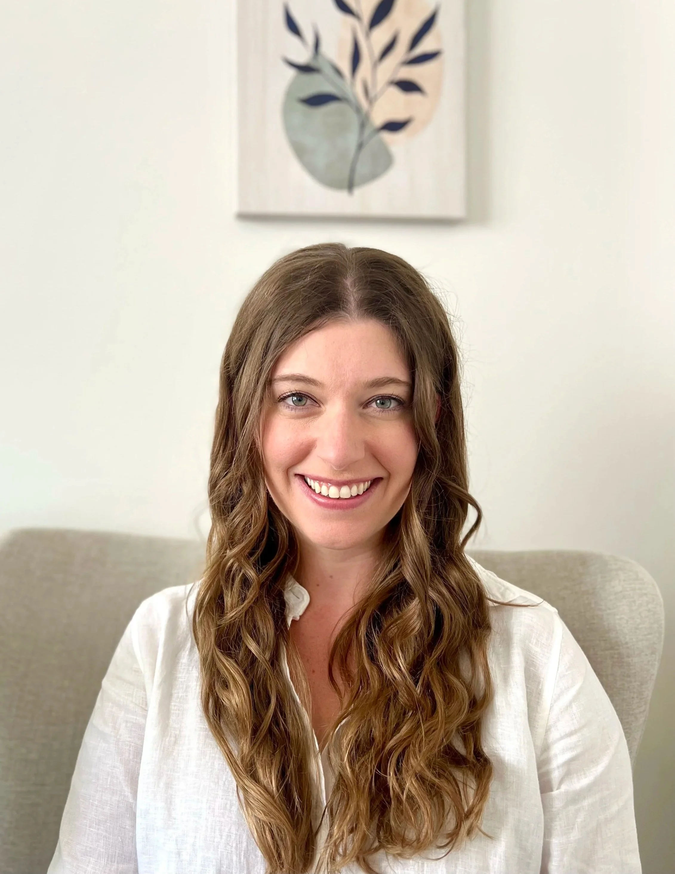 A woman with long wavy brown hair is smiling and sitting on a beige chair with a framed abstract leaf artwork on a white wall behind her.