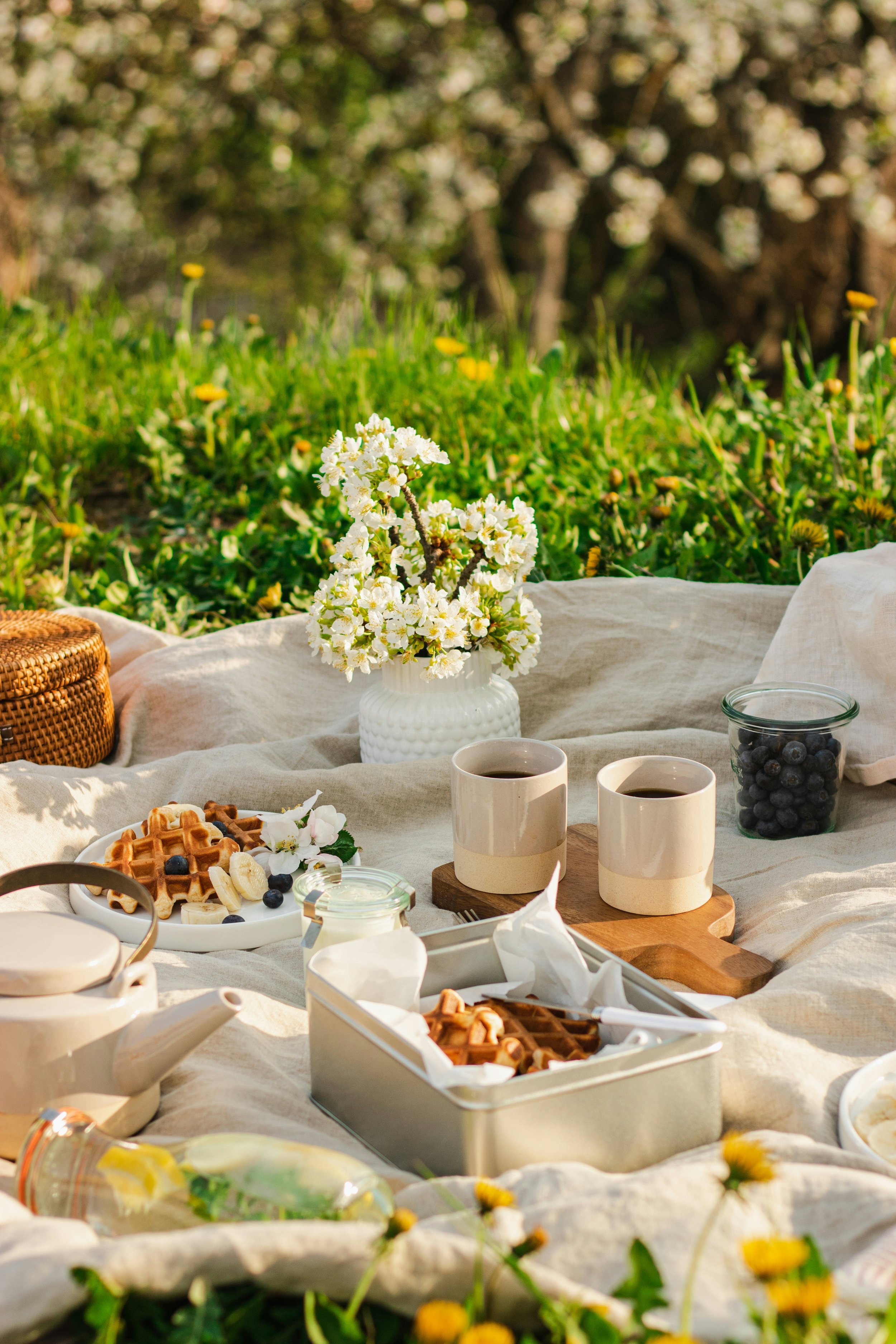 A picnic setup on a beige blanket in a lush green outdoor area with blooming trees. It includes a white vase with white flowers, cups of coffee, a jar of blueberries, waffles with fruit, a teapot, and other breakfast items.