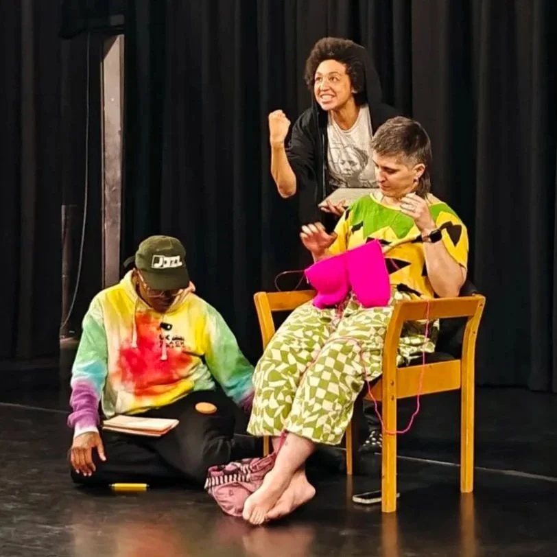 Amelia sat knitting, Adae on the floor reading, Charmaine behind smiling and pumping her fist.