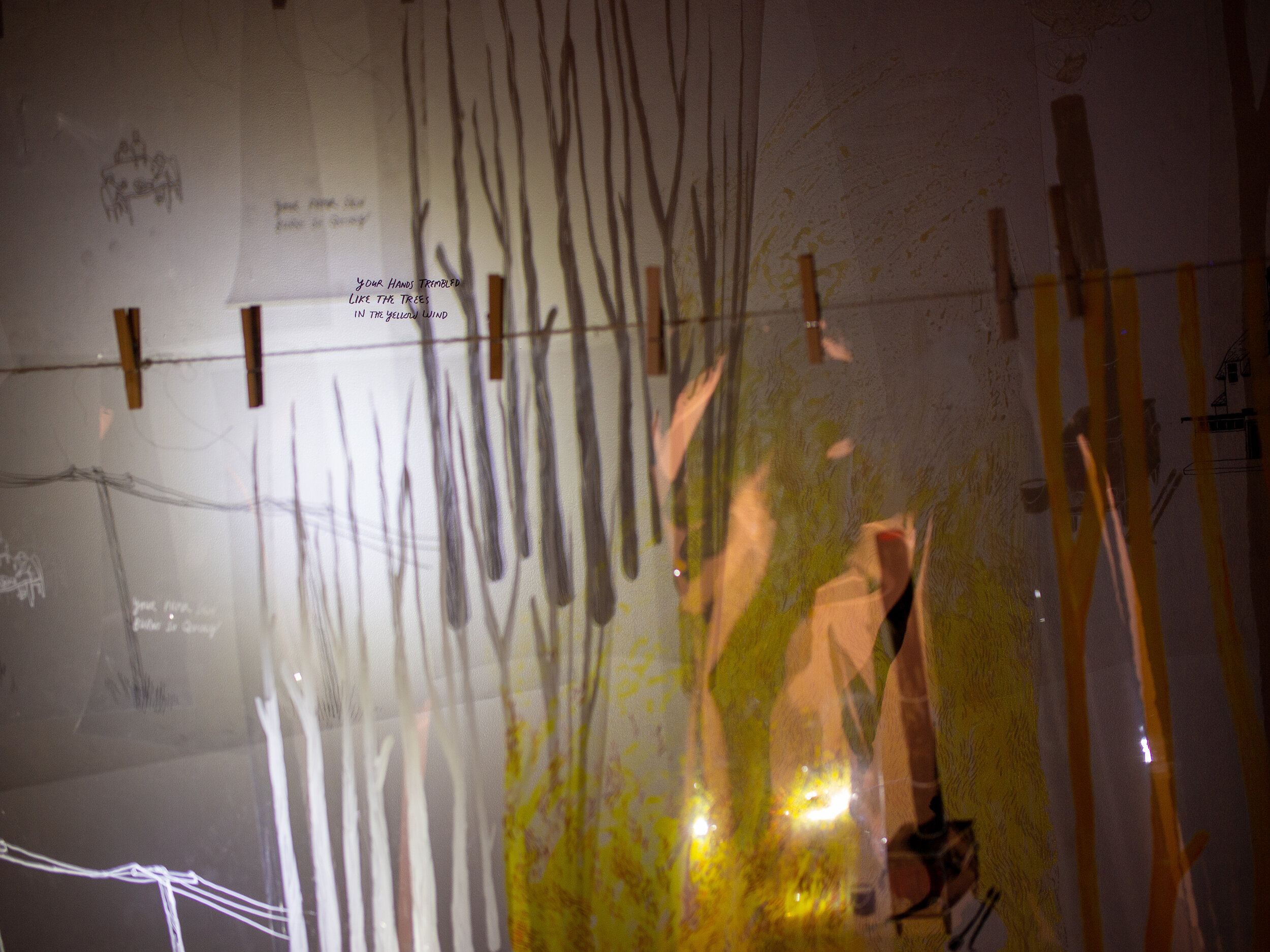  Clear plastic sheets with drawings on them hang on a clothesline, with a white light shining through them to create shadows of the drawings on the wall behind it. There are also images and text that have been put on the wall directly. We are too clo