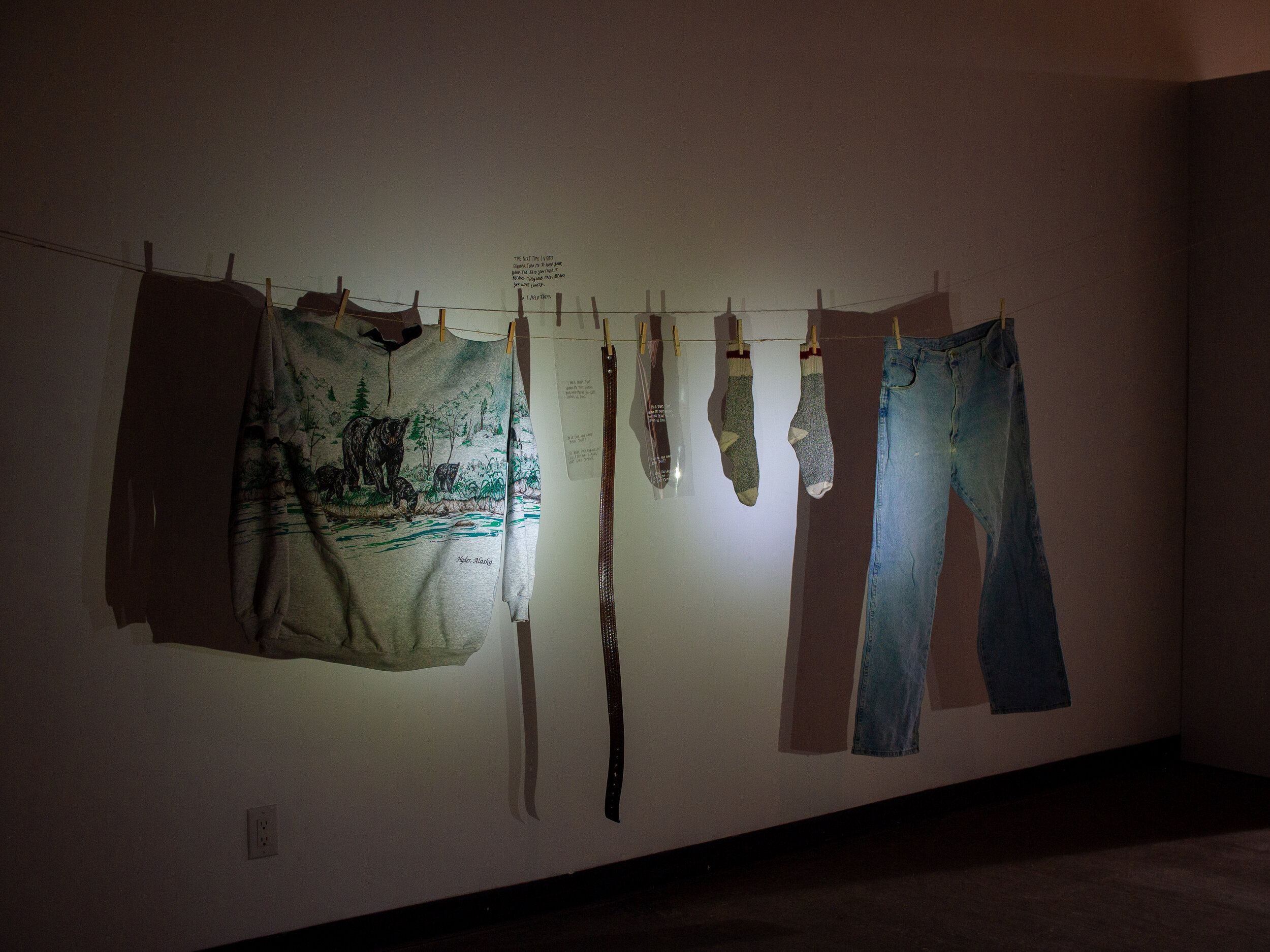  An outfit’s worth of clothes hang on a clothesline in a darkened room. A white light shines on them, making shadows of each item on the wall behind the clothesline. From our left to our right, there is a grey sweater with a drawing of a bear and its