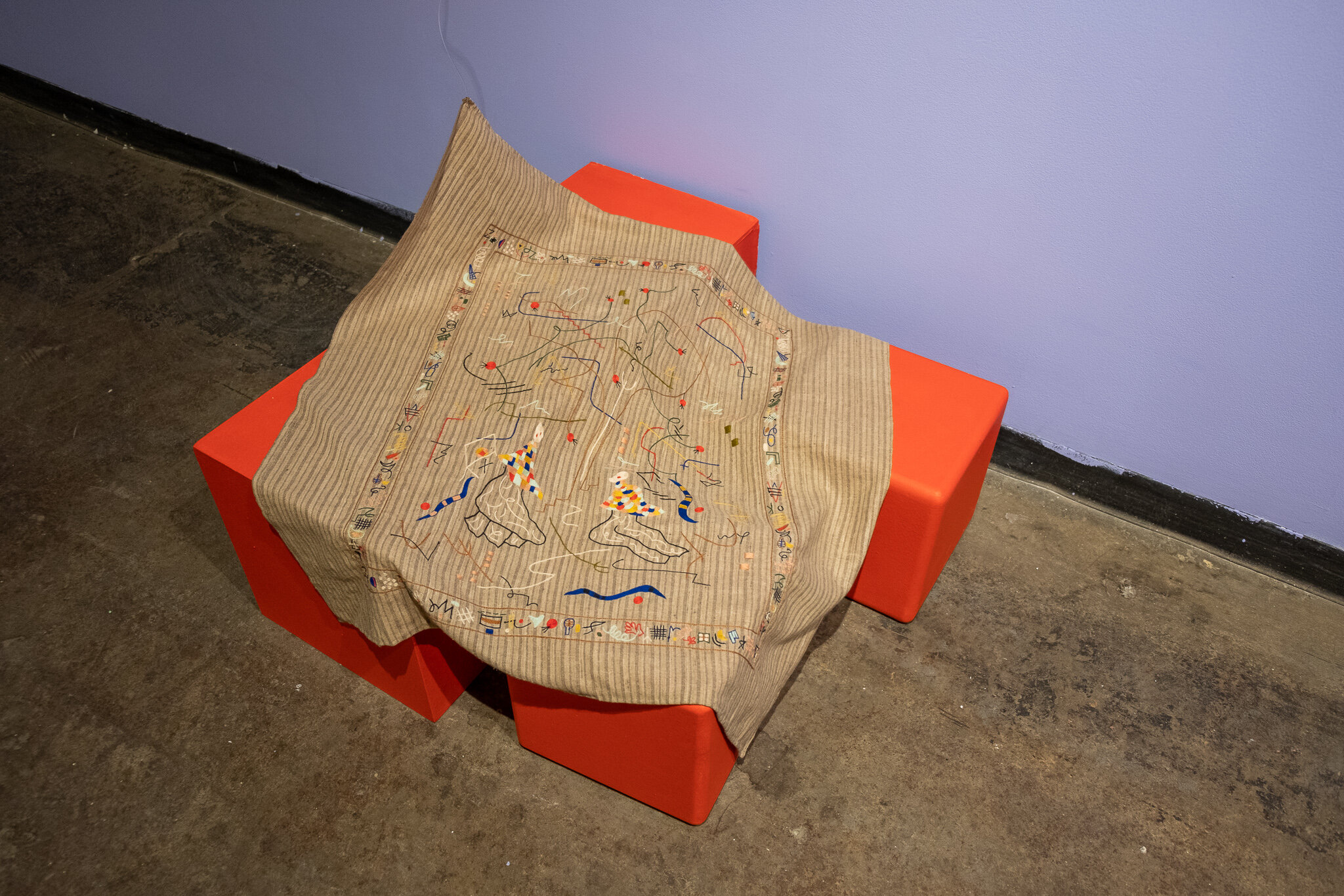  This is a brown and tan striped piece of fabric that is laid across three bright red plinths, two of them being small cubes and one of them being a rectangular box. On the fabric there is embroidery work, consisting mostly of curved, abstract lines 