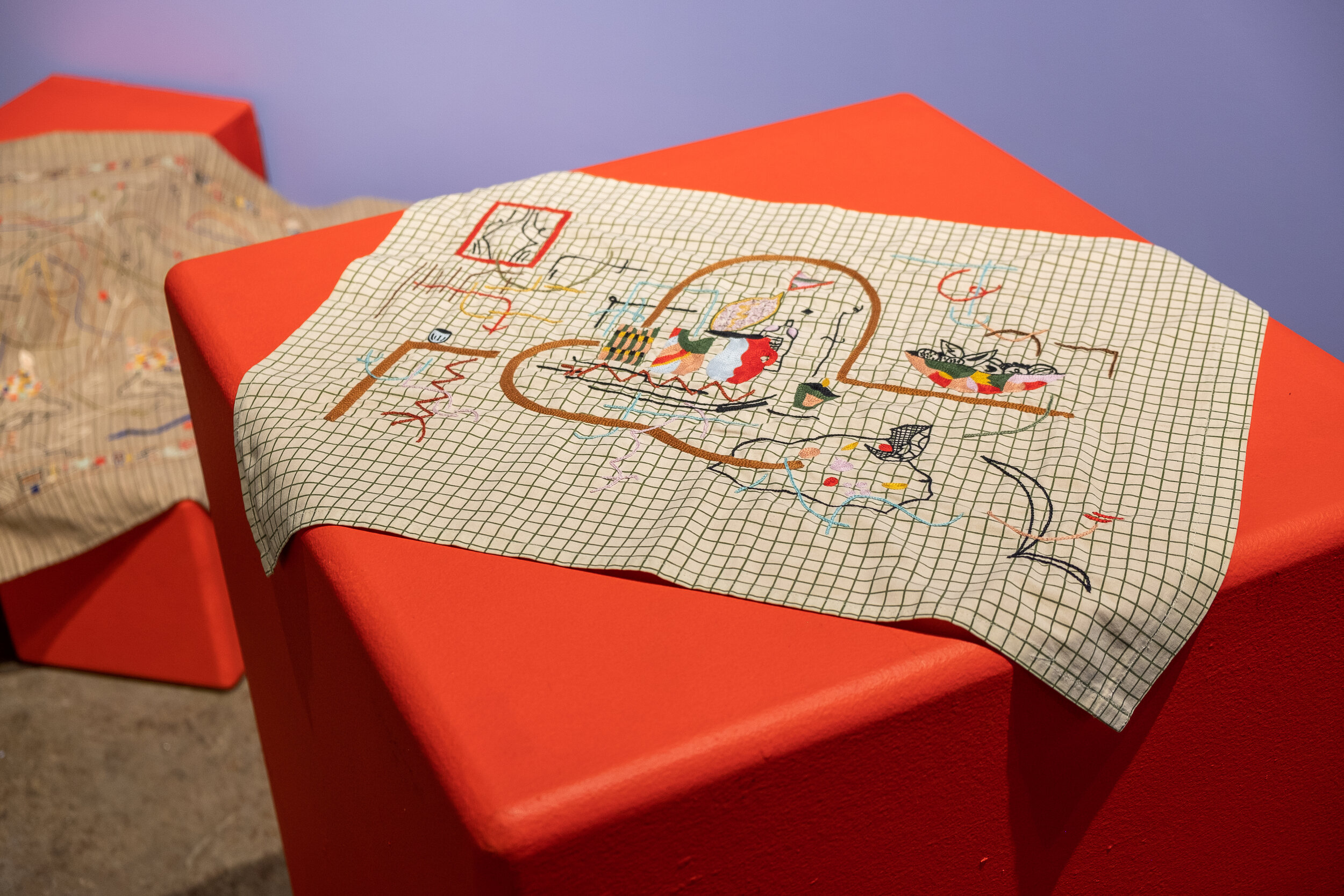  This is a piece of white fabric with a black grid design laying on a bright red plinth, with the corners falling off of the edge. The fabric is embroidered with what looks to be a sort of still-life scene. Moving from left to right there is a red wi