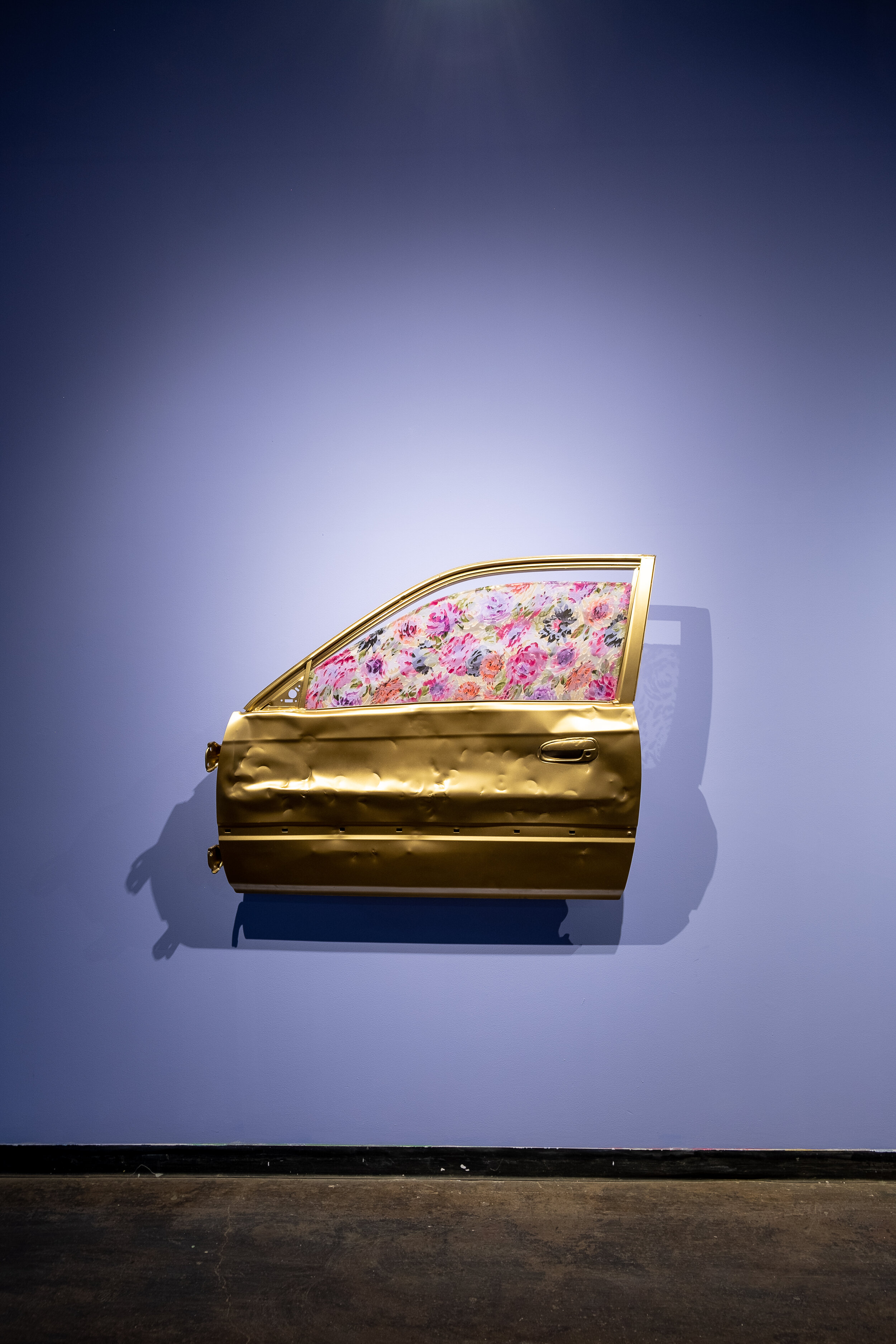  A golden car door is hung on the wall. The door has been completely spray-painted gold, and the door has multiple large dents in it. The window does not have clear glass, but rather a window covering decorated with pink, purple, and orange flowers. 