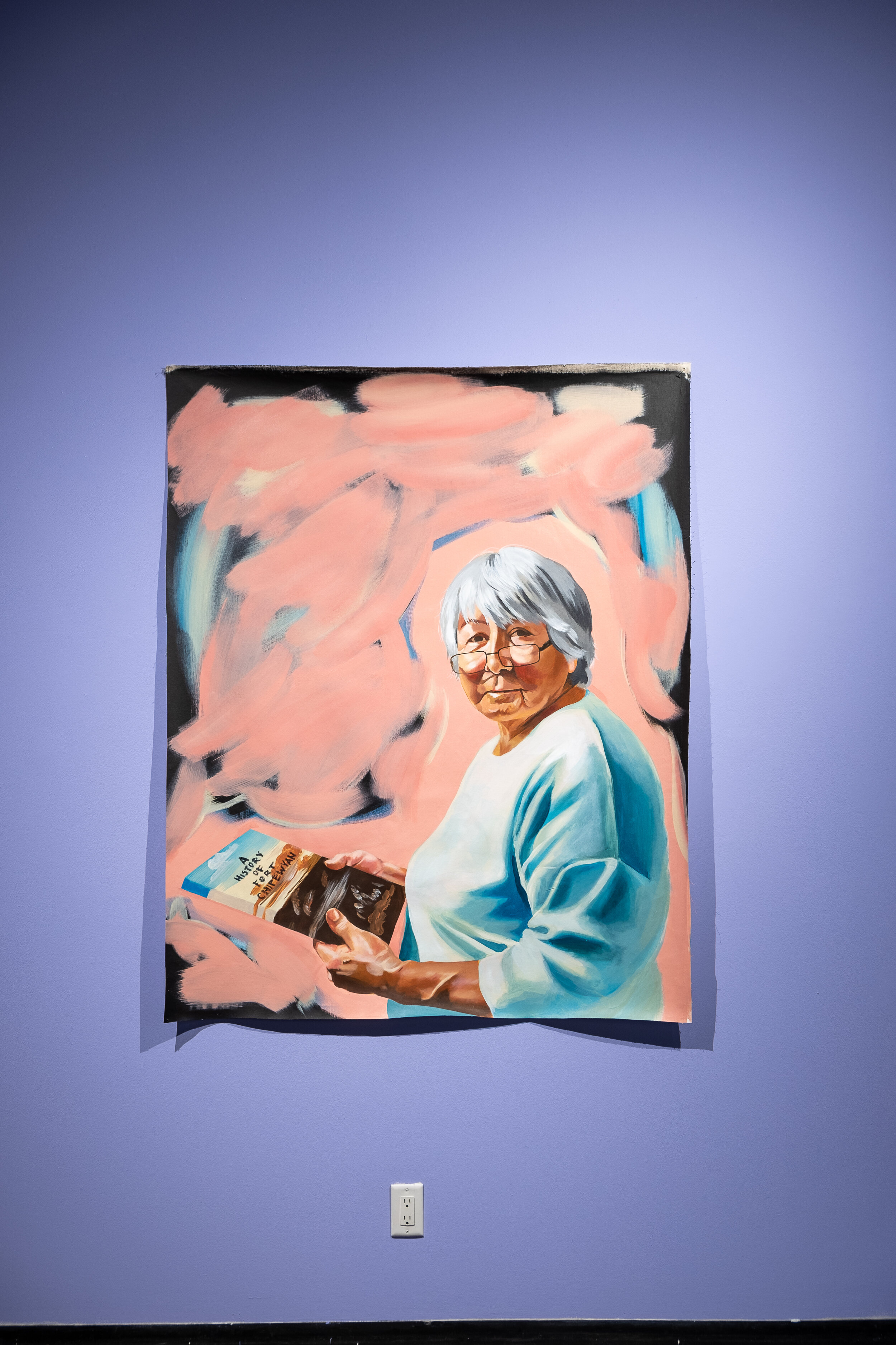  A painting of an old woman with warm, tanned skin, light grey hair, dark eyes, and a rounded face. The woman wears a long-sleeved, light blue shirt, and thin, black square glasses. Her body is turned away from us to the left of the painting but she 