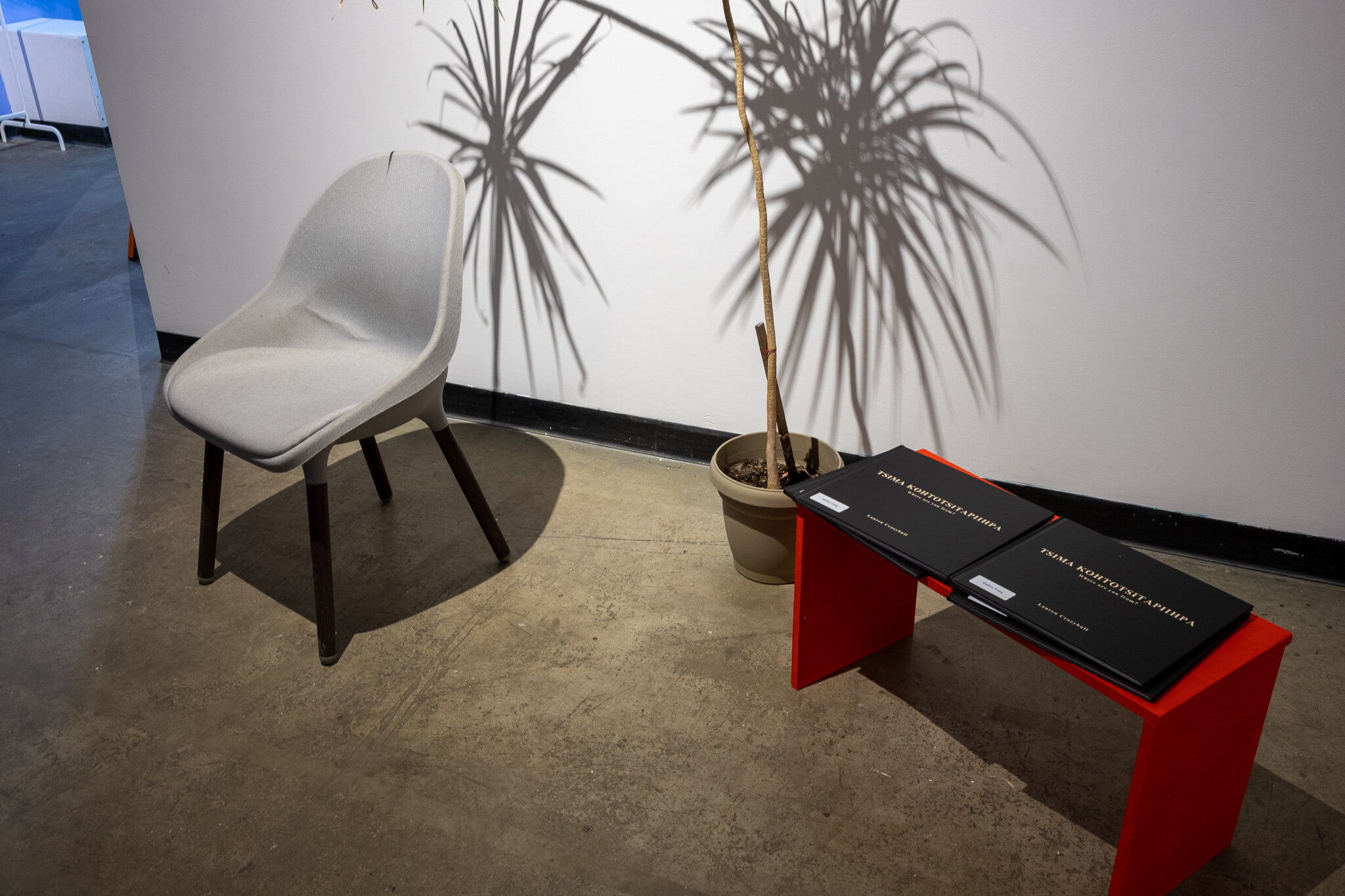  A picture of a short red table and chair in the gallery space. The table is holding wide, black exhibition books with the title “TSIMA KOHTOTSITAPIIHPA” followed by the subtitle “Where are you From?”. The name “Lauren Crazybull” is at the bottom of 