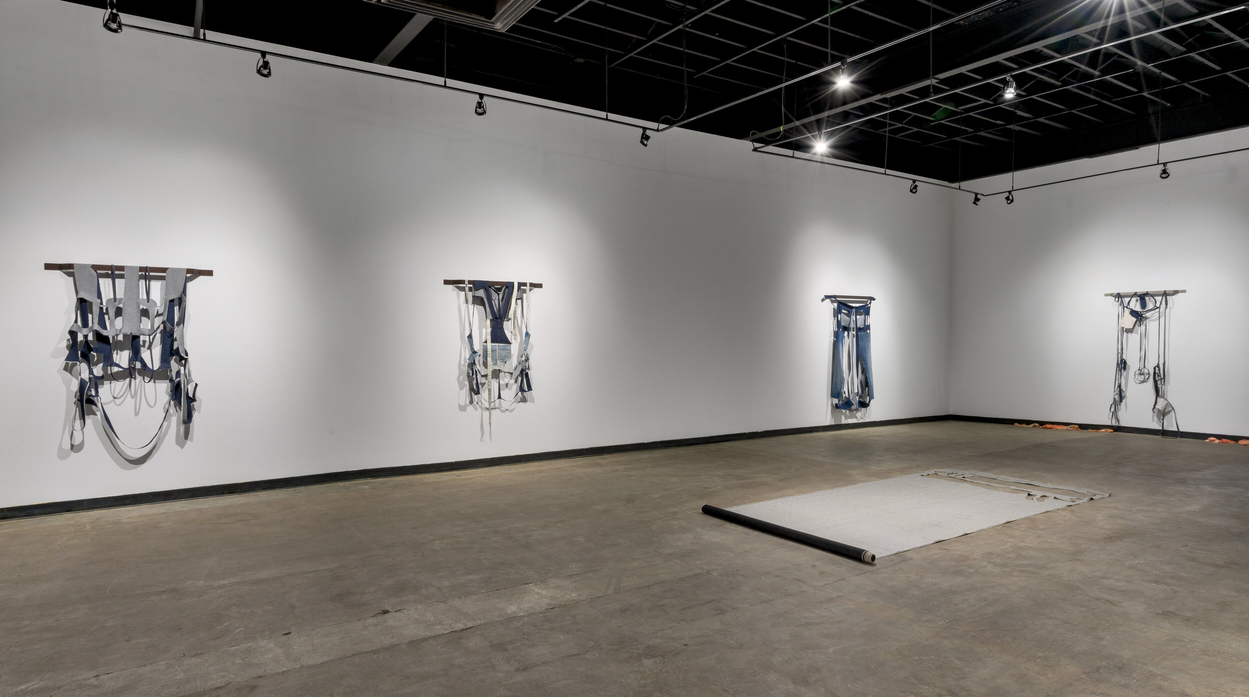  This is a wide view of the gallery for Karen Kraven’s exhibition, Lull. There are four hanging works on the wall, which look like various denim clothing items that have been deconstructed. There is also a piece that has been rolled out on the floor,
