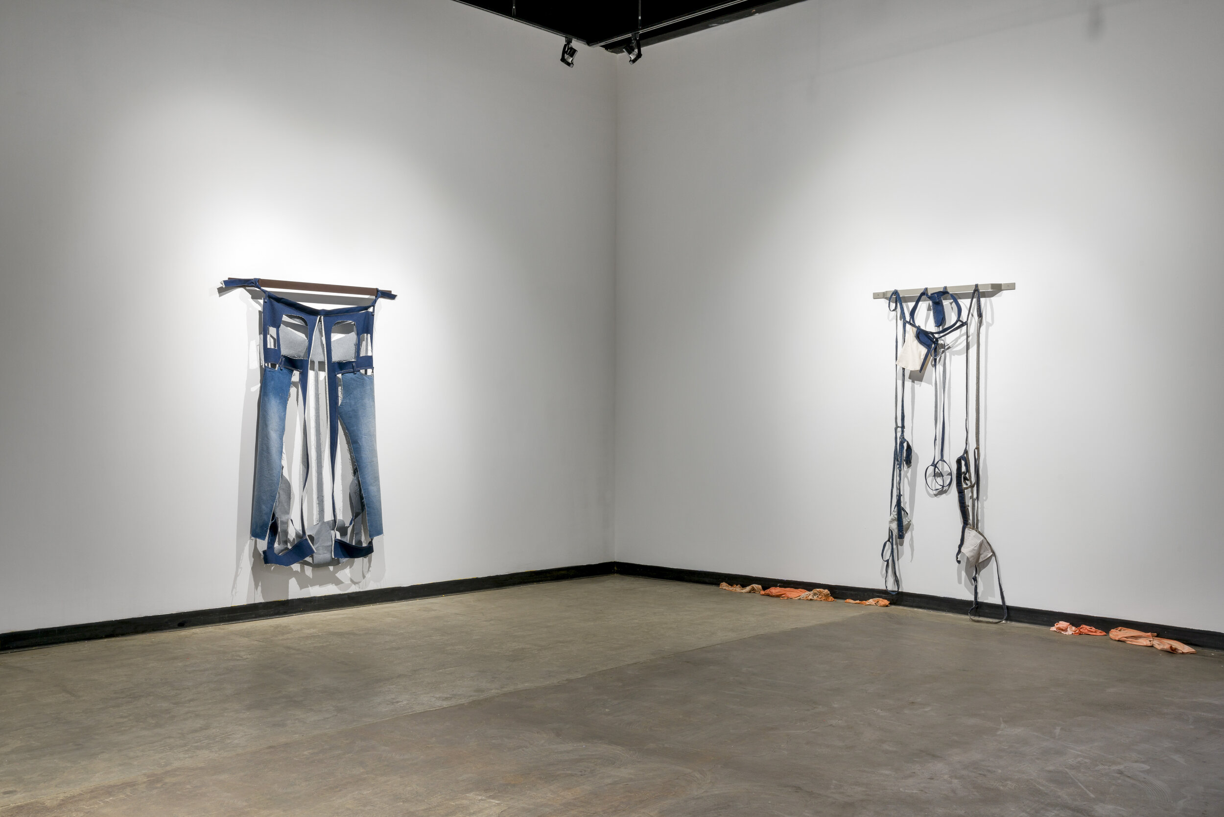  A look at two pieces as they hang on two walls that meet in a corner. The piece on the left wall looks similar to a pair of blue jeans, if you had cut out the back pockets, creating square holes in the denim. The piece on the right is primarily long
