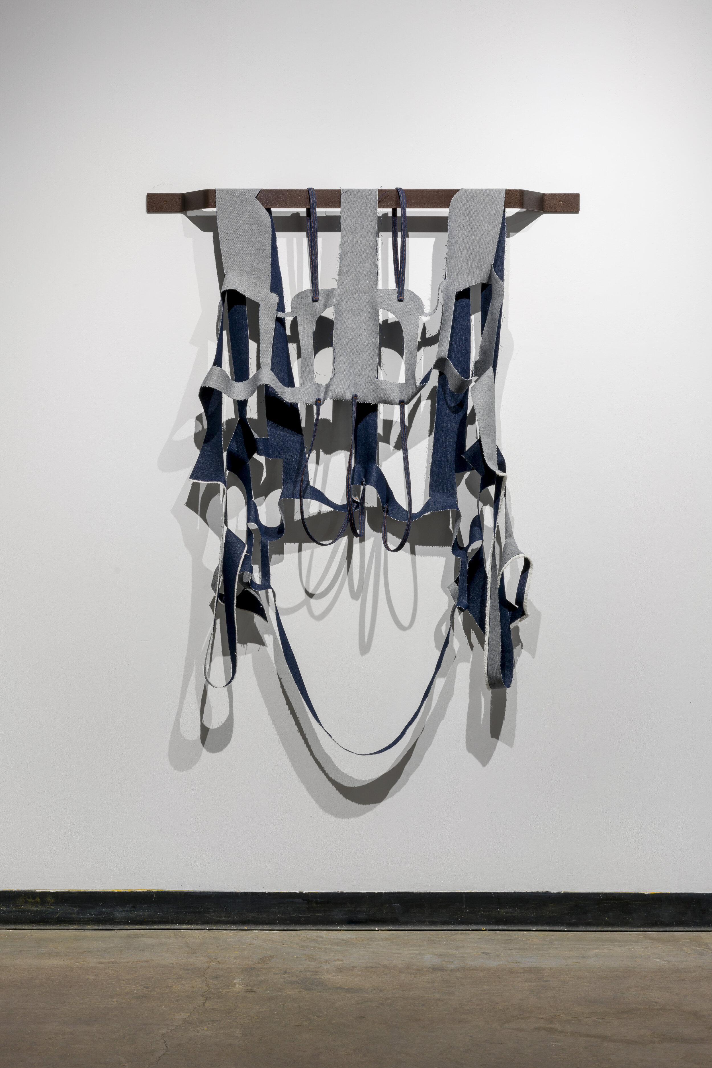  A piece of fabric hangs from a square metal rod attached to the wall behind it. The fabric looks as if a pair of dark blue jeans have been cut apart, making an abstract shape. The top of the hanging fabric has thicker cut pieces, making blocky recta