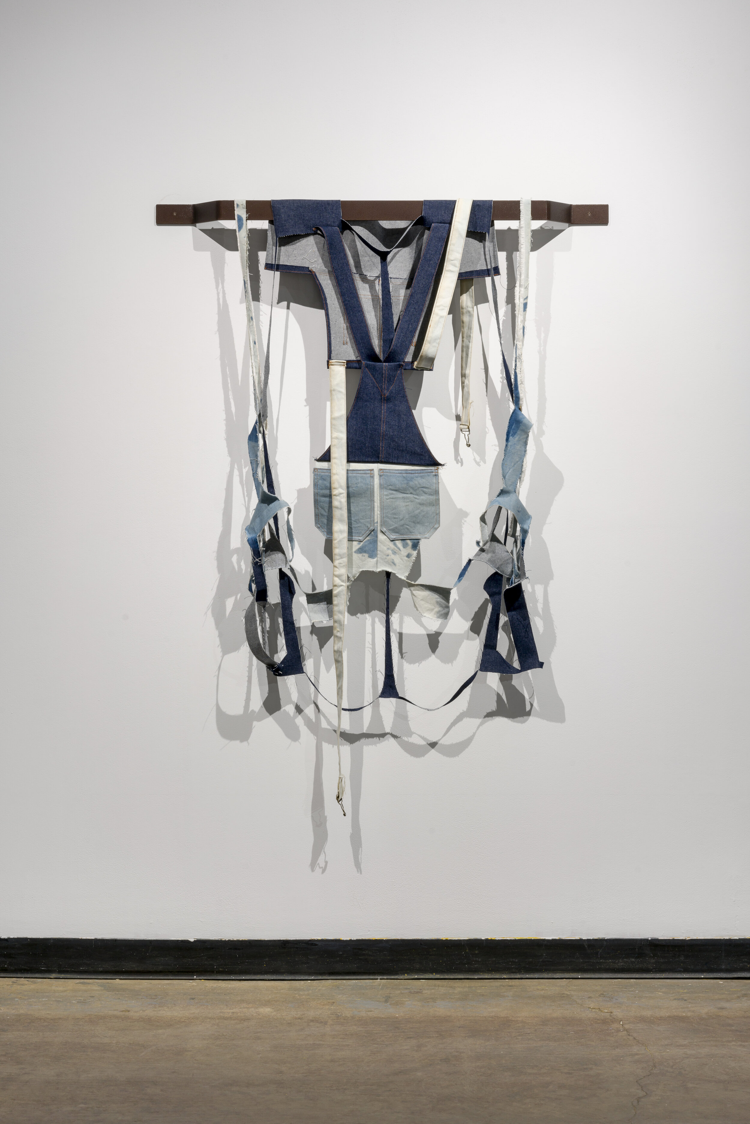  This is a hanging piece that is made of a combination of dark blue, light blue, and white denim. The central piece of fabric looks like the back of a pair of overalls, where the shoulder straps come together. On both sides of the central piece, ther