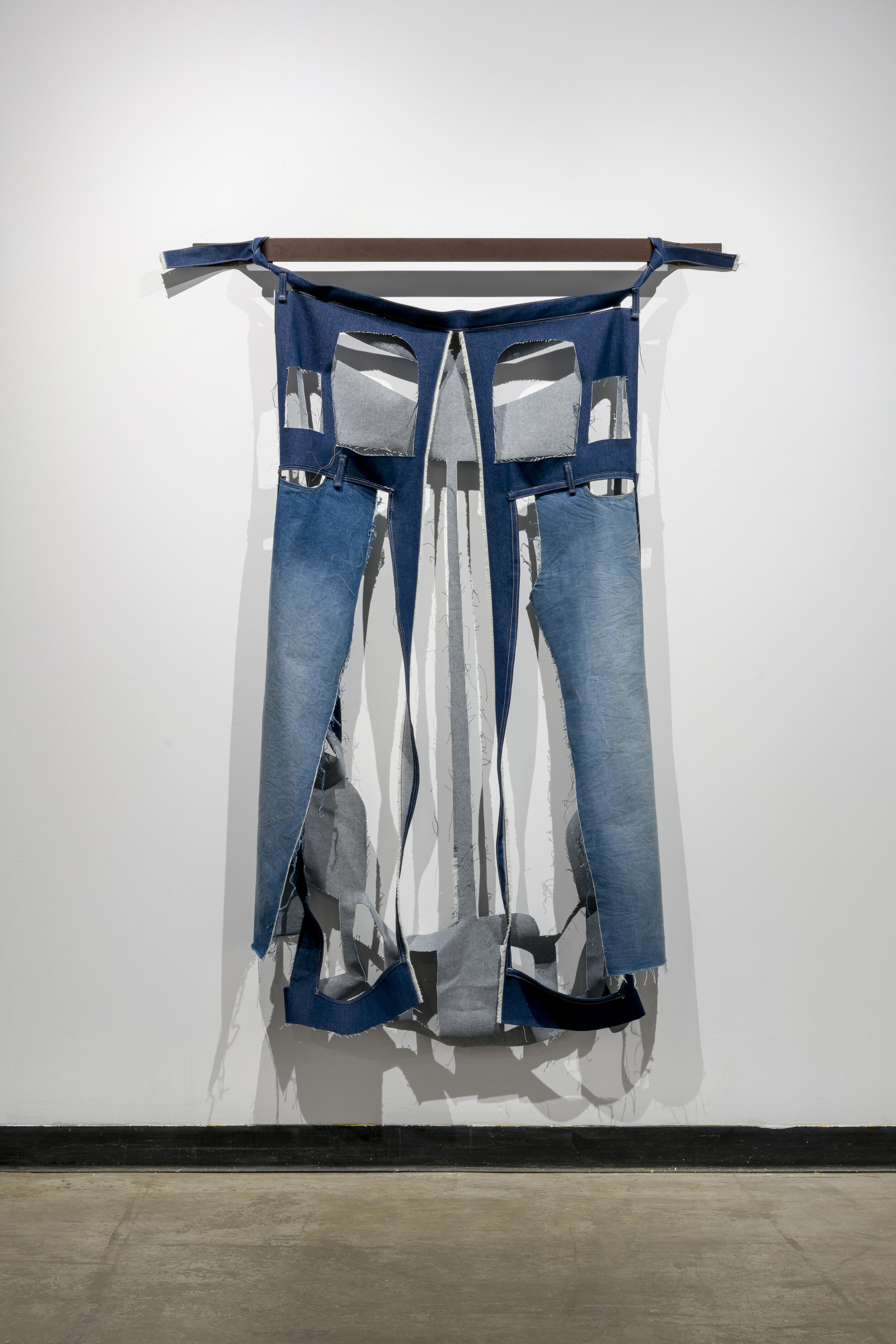  This is a hanging piece that looks to be made of a pair of blue jeans that have been deconstructed. This piece resembles a pair of jeans in shape, but notably squares have been cut out in the area where the pockets would be. The pants also look to h