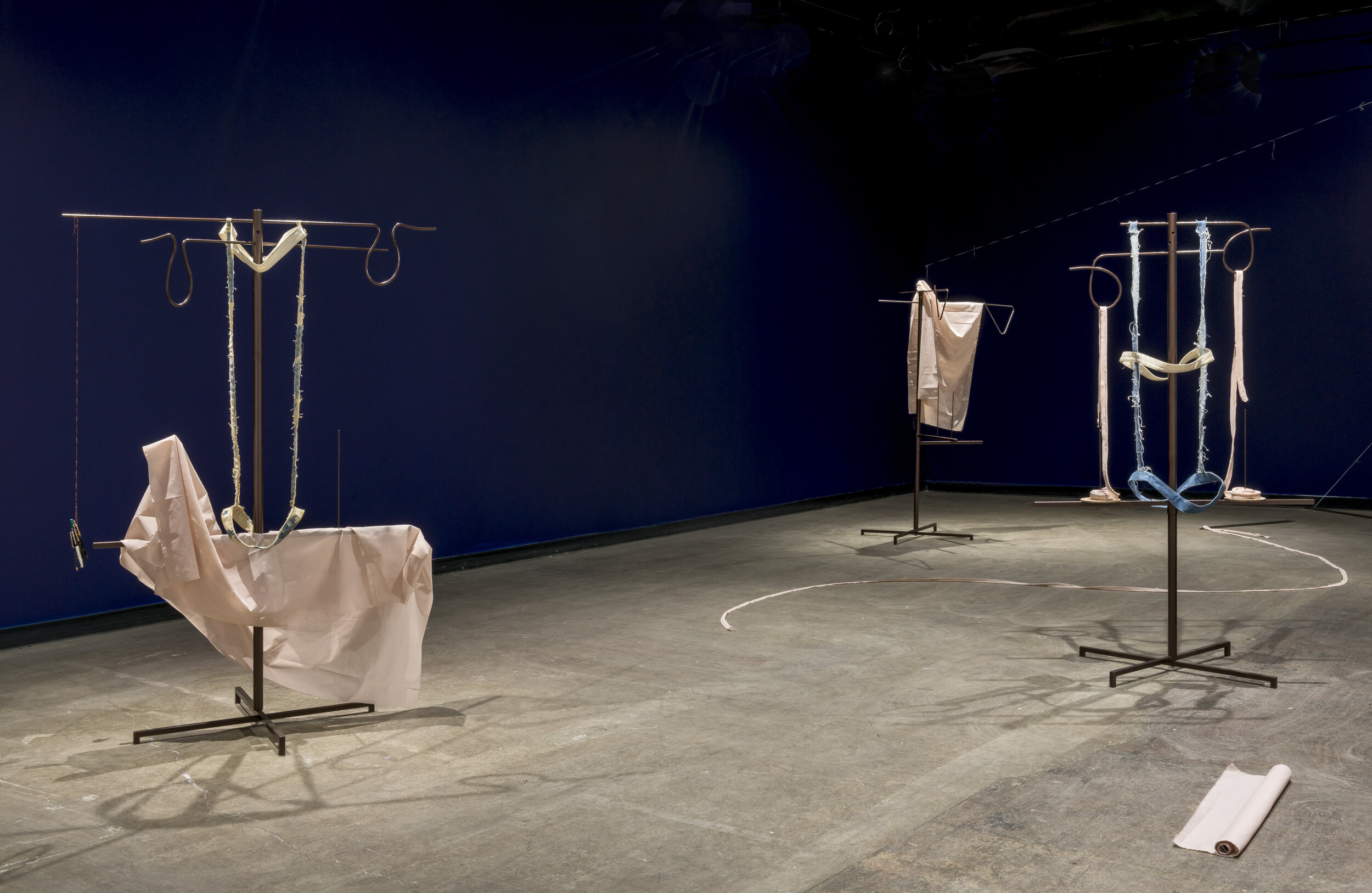  This is a wide view of the gallery for Karen Kraven’s exhibition, Lull. In this room, there are three free-standing structures. Each of these structures is a metal pole on a stand with a couple bars running through them, and pieces of fabric hanging