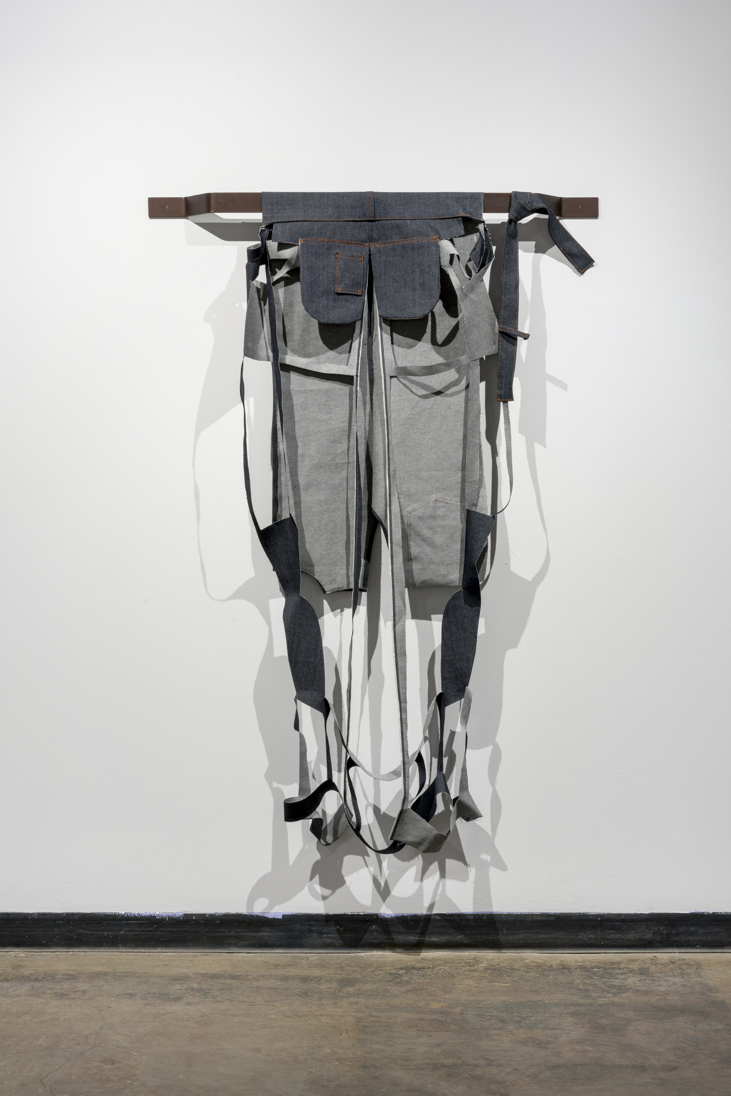  This is a hanging piece made by deconstructing a pair of grey jeans. Most of the pant legs have been cut away, leaving outlines of them through thin strips of fabric. What seems to be intact from the jeans in this piece are the waistband, the back p