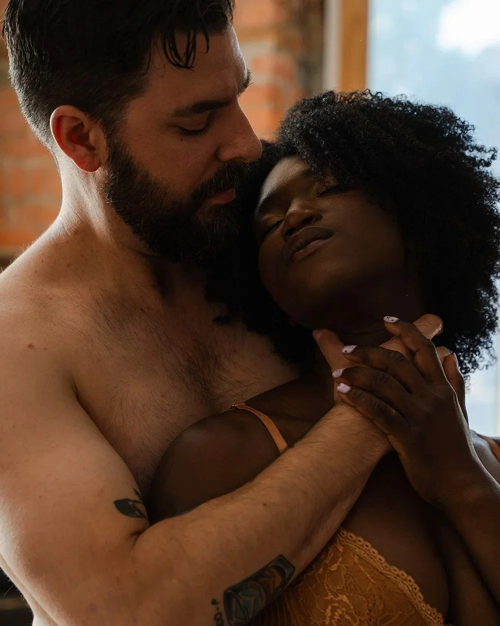 Boudoir photography in Hamilton with shirtless man with a beard gently holds and leans toward a woman with curly hair, who is relaxing and has her eyes closed, in a cozy indoor setting.