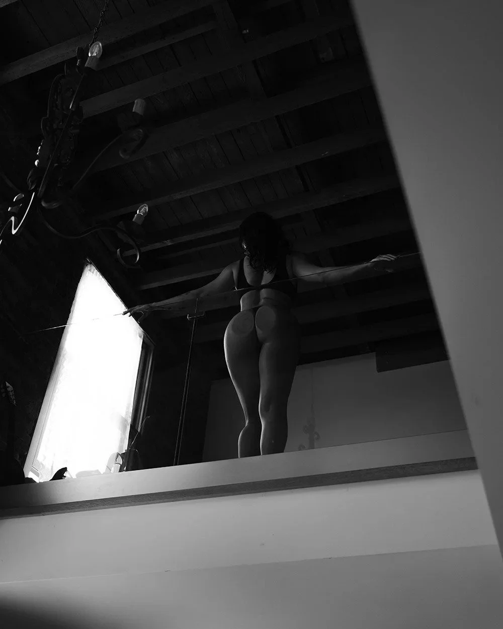 Hamilton boudoir photography with woman in sensual lingerie standing on a mirror platform, stretching with arms extended, in a dimly lit room with wooden ceiling beams and a large window letting in natural light.