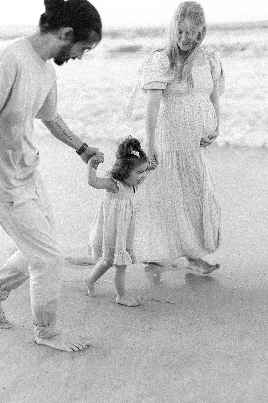 A family of three, including a man, woman, and young girl, holding hands and walking barefoot on the beach. Taken by Wylde Folk Studio, Brisbane. Maternity photography.